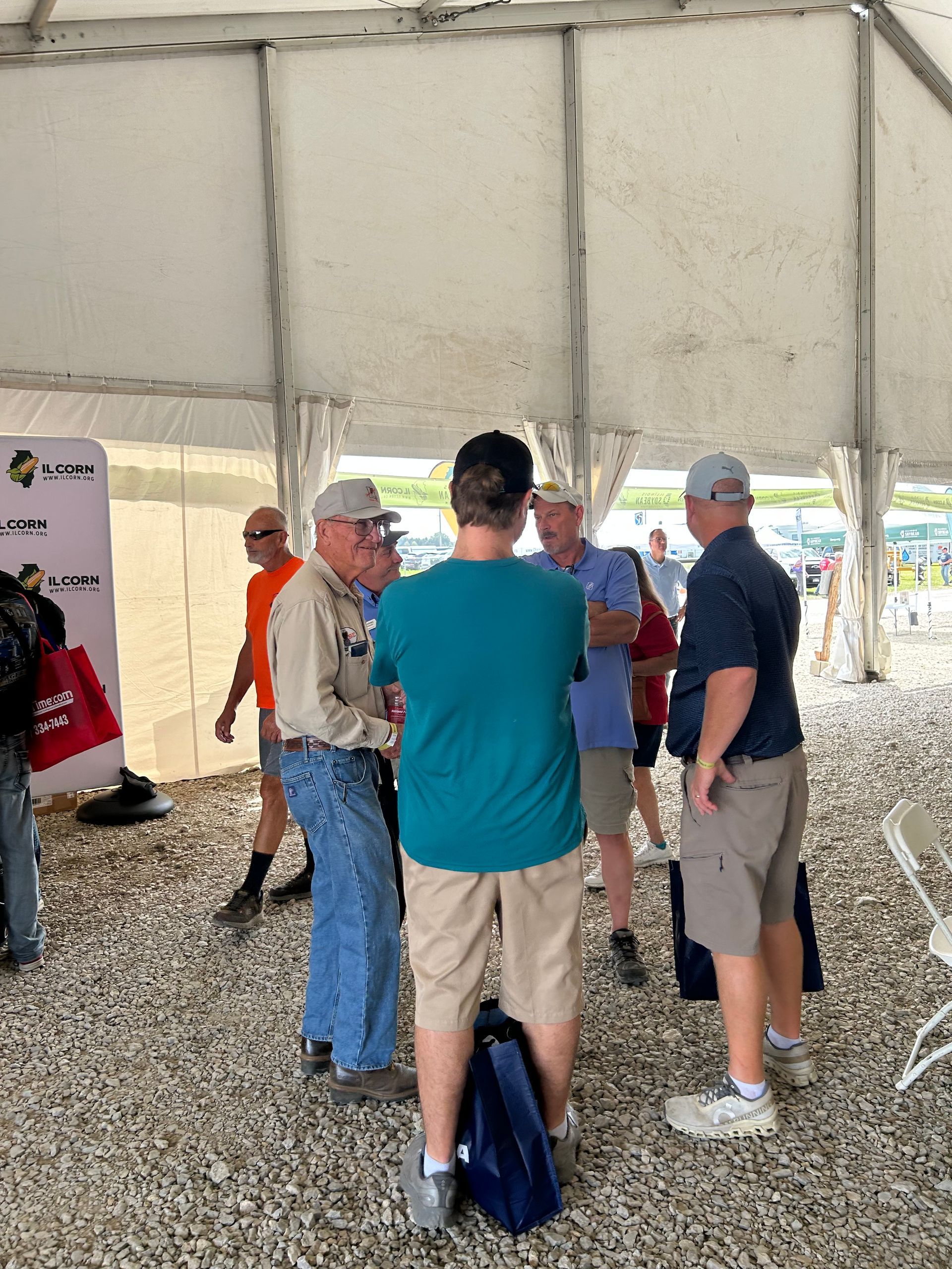 People in a tent, talking. Some wearing hats, a mix of casual attire. Tent floor is gravel.