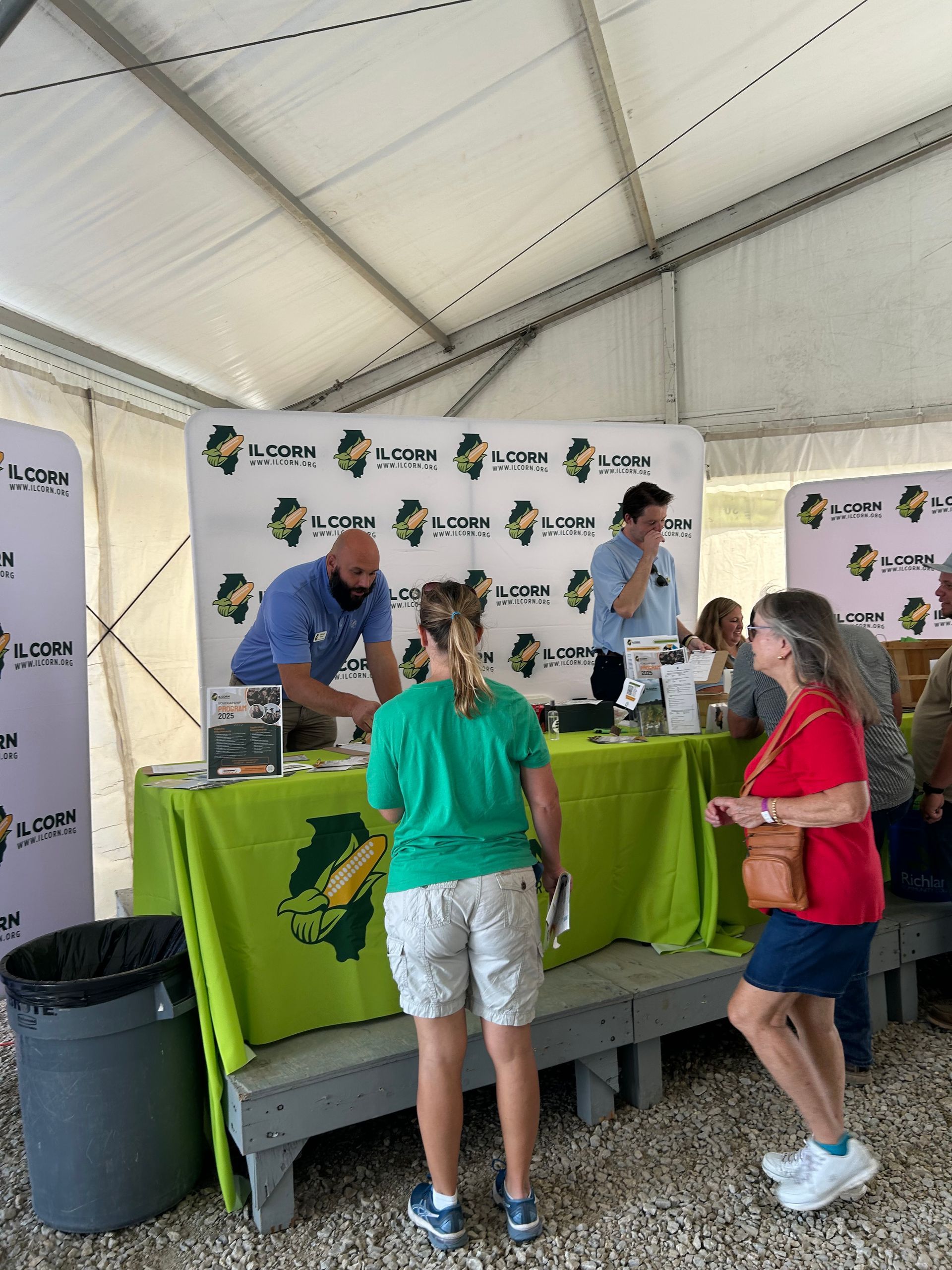 People at a booth with green and yellow signage under a tent. Two men stand behind a table, interacting with visitors.