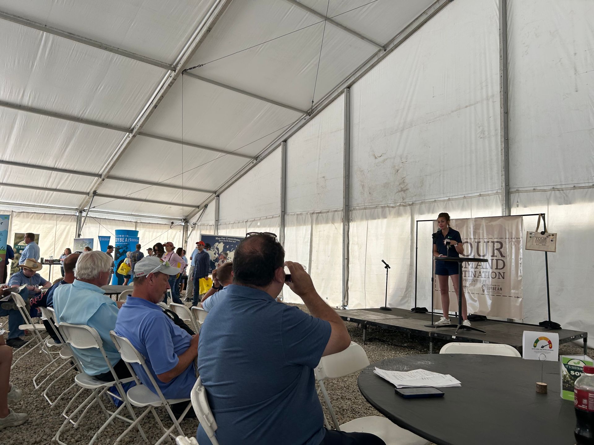 A person speaking on a stage inside a tent to an audience seated in folding chairs.