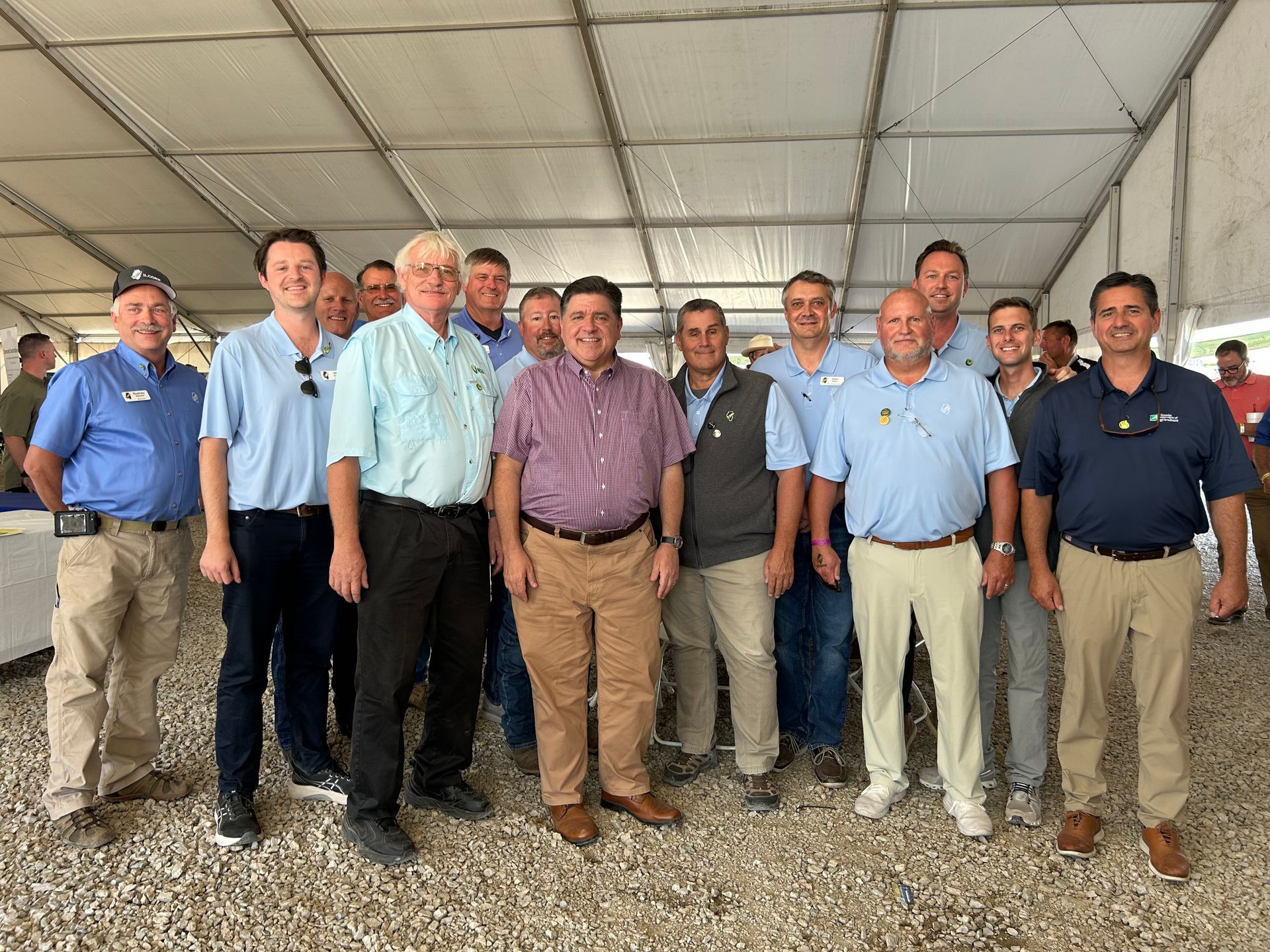 Group of men posing under a white tent. Several wear blue or khaki shirts.