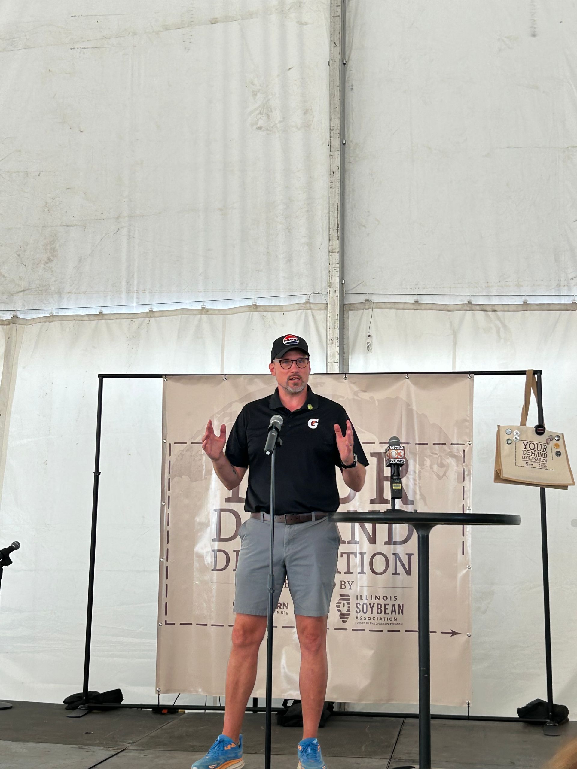 Man speaking at a podium. He's wearing a black shirt and shorts. The backdrop has a banner and a bag.
