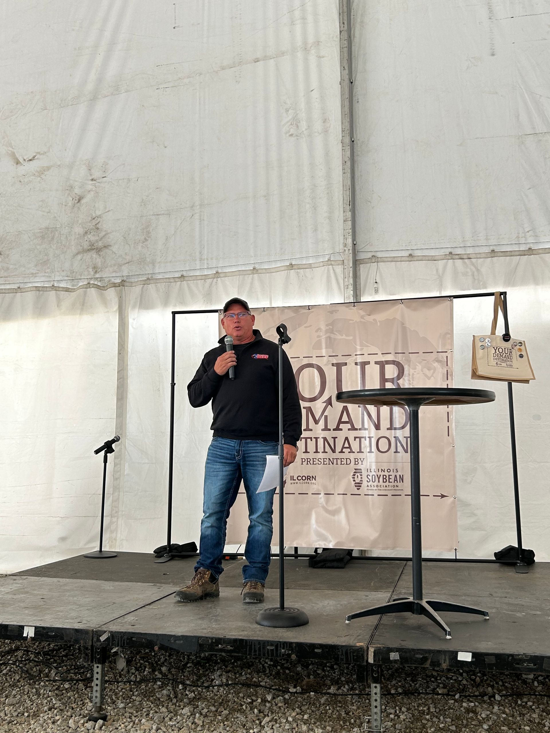 Man speaking at podium under a tent. Brown hair, dark sweater, jeans, outdoor setting. Backdrop: