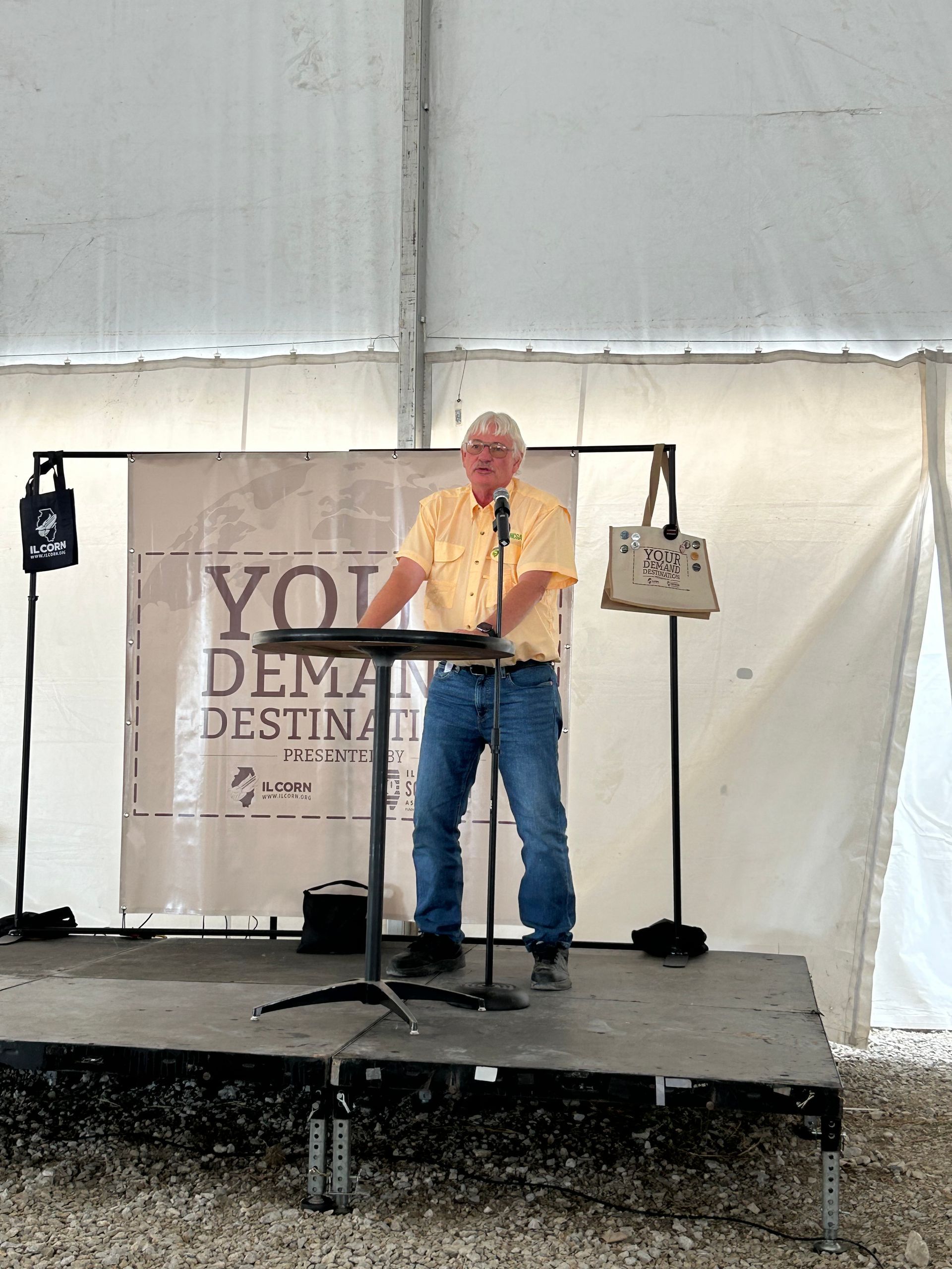 Man speaking at a podium on a stage in a tent, banner behind him.