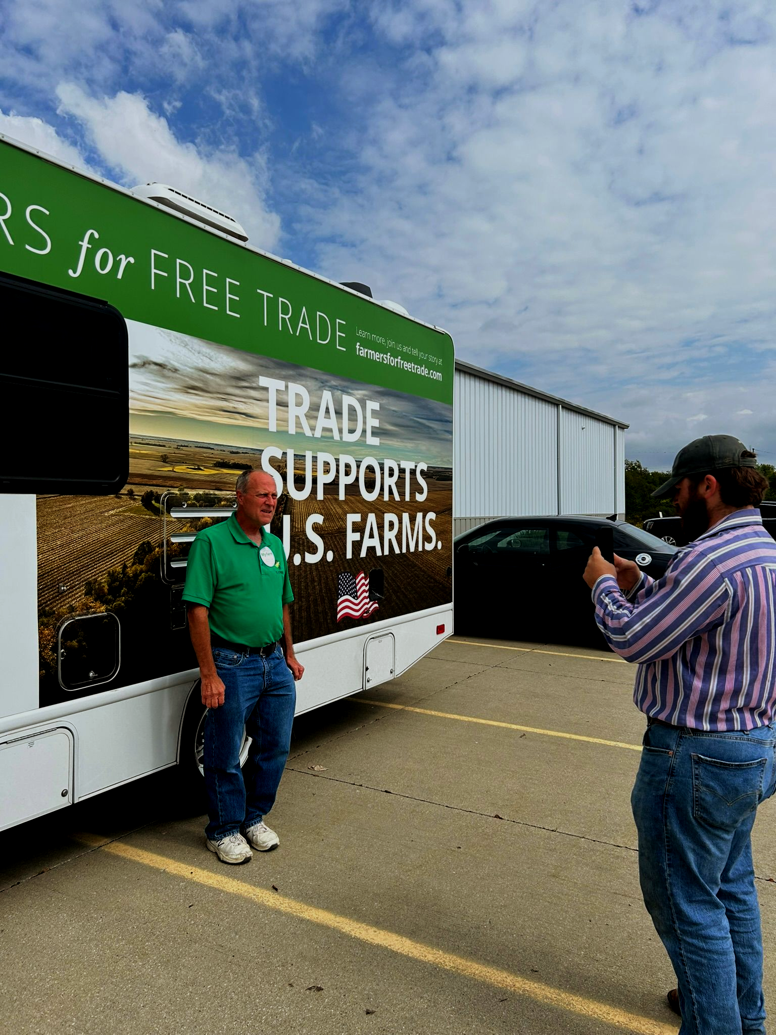 Man poses in front of a truck promoting free trade; another man takes his photo. Blue sky, green shirt, jeans.