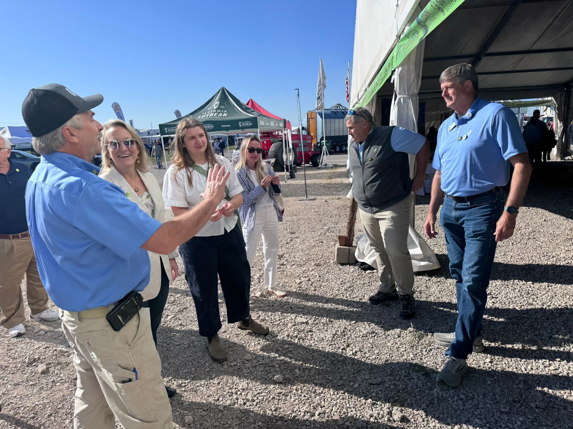 Group of people outdoors; man in blue shirt gesturing, others listening, tents in background.