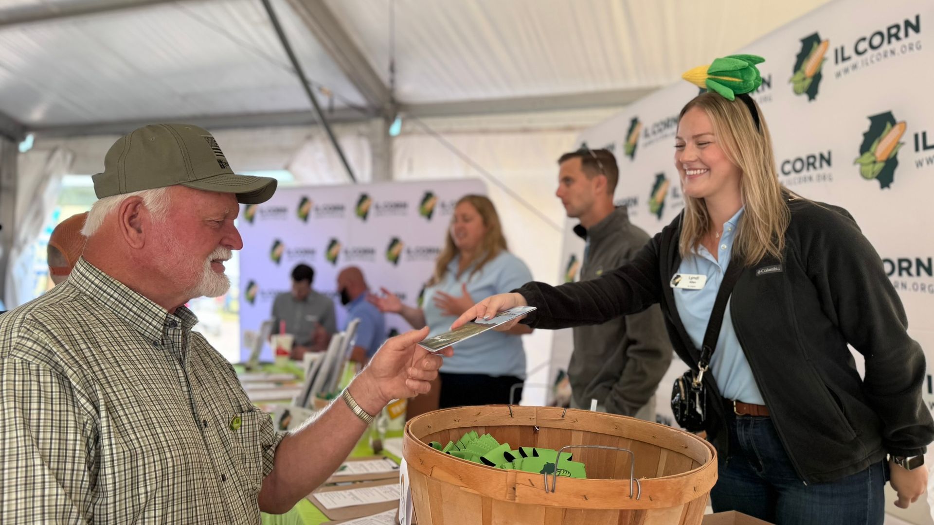 A woman in an ear of corn headband hands something to a man at an IL corn event.