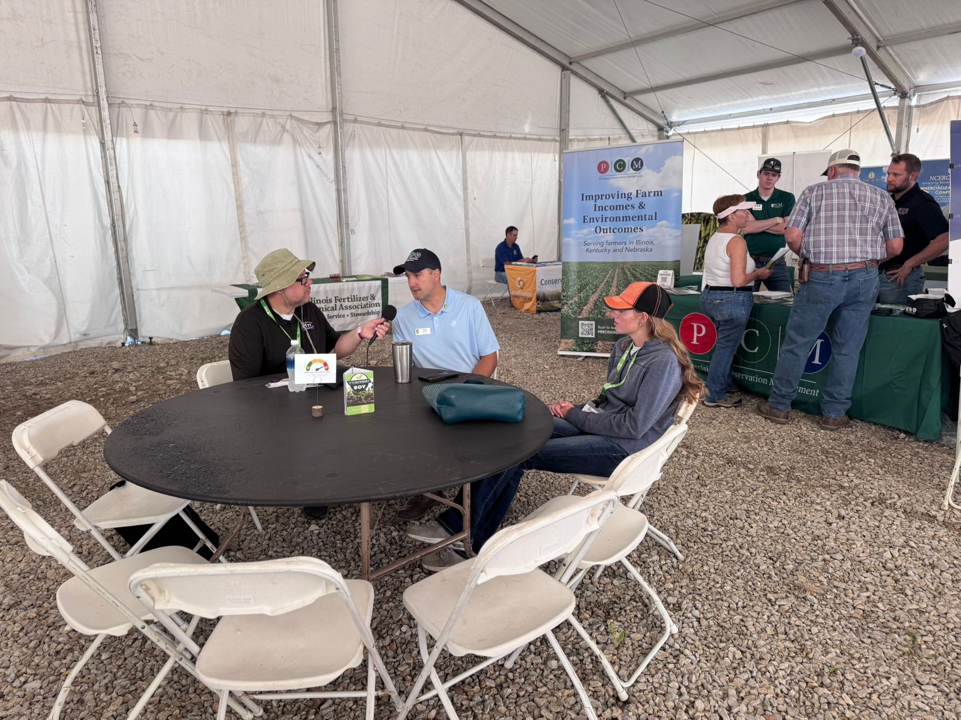 People at a trade show, sitting and standing, talking near a table inside a tent.