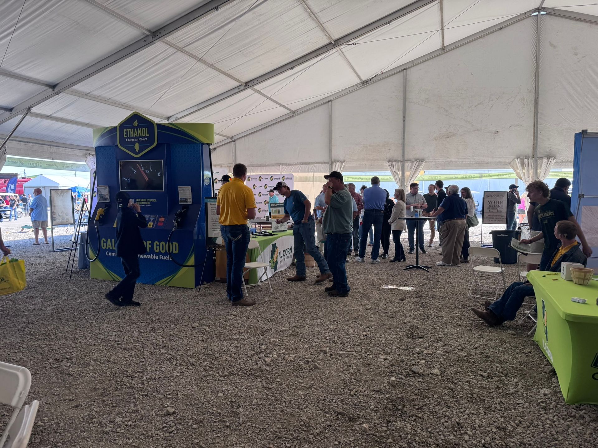 People browse booths at an outdoor event under a tent. The floor is gravel.