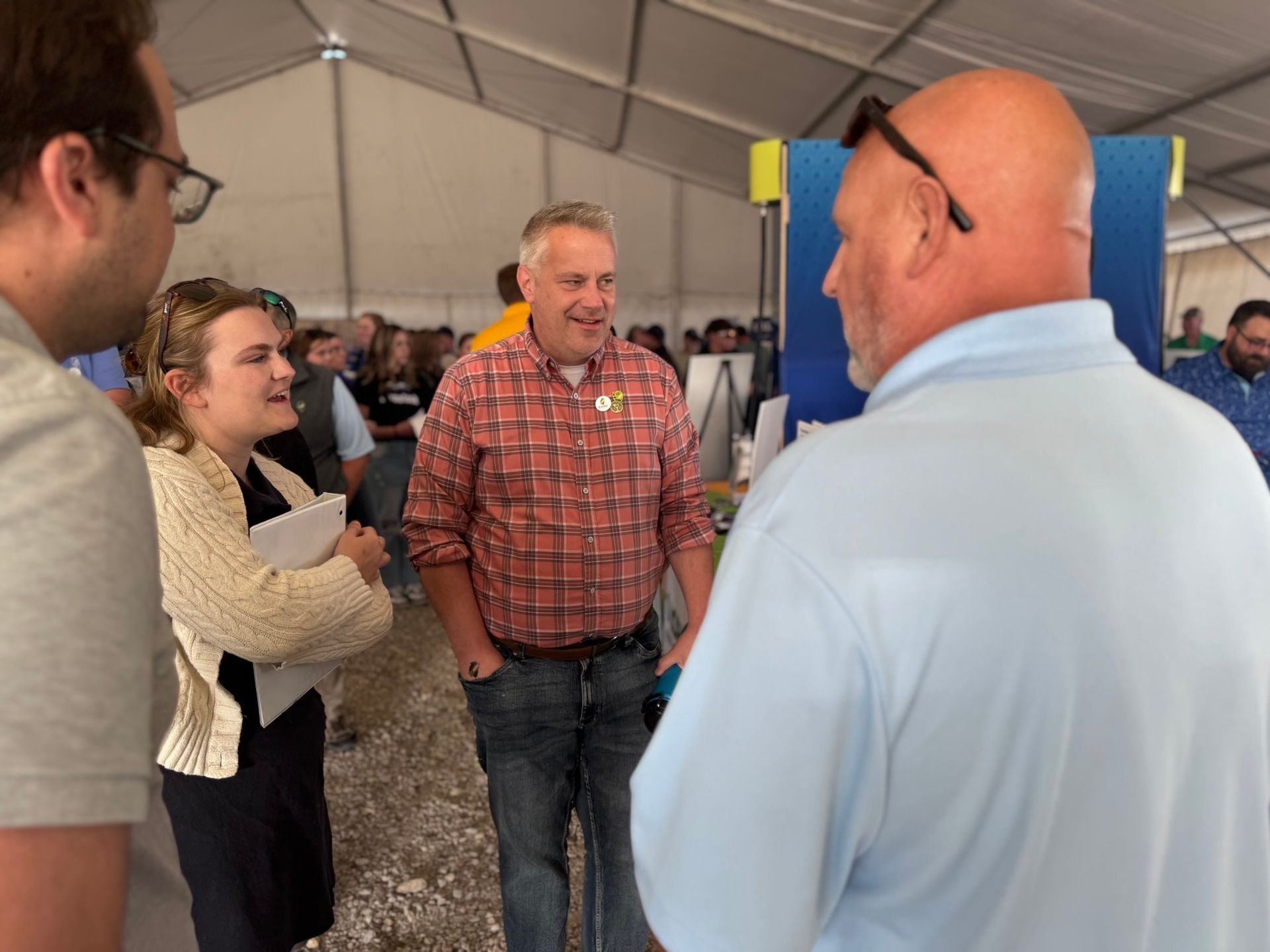People in a tent, conversing. Man in plaid shirt smiles, others listen. Setting appears to be an event.