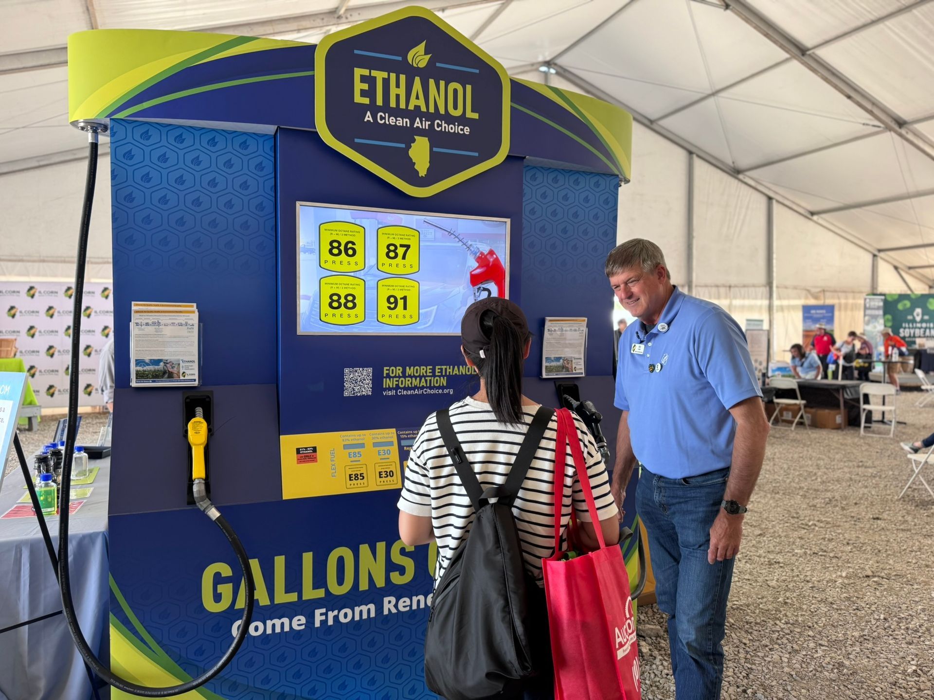 A man and woman at an ethanol exhibit. Blue and yellow display, digital screen showing fuel options.