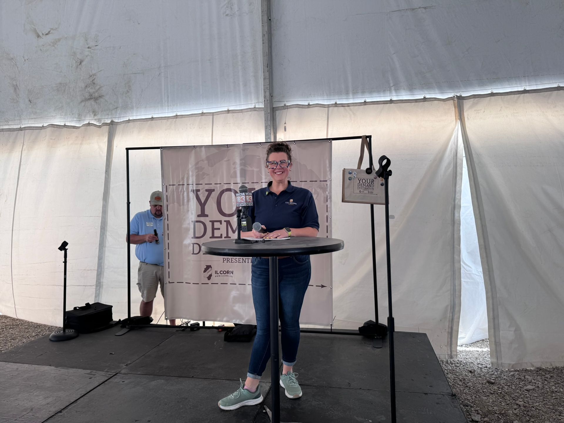 Woman speaking at a podium, wearing glasses, in a tent. A man stands in the background.