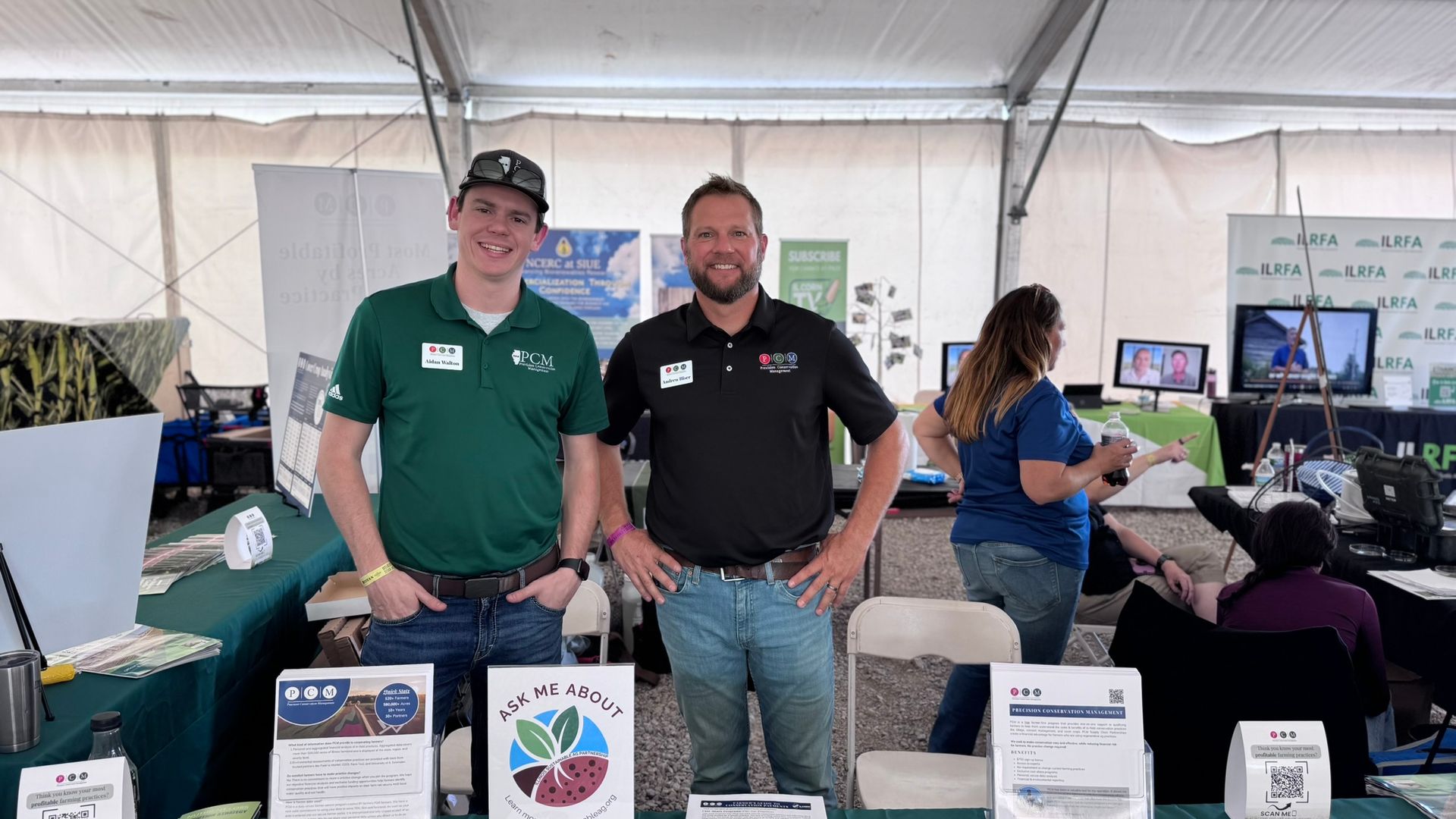 Two men at a trade show booth, smiling. Tables with displays, people in background. Inside a tent.