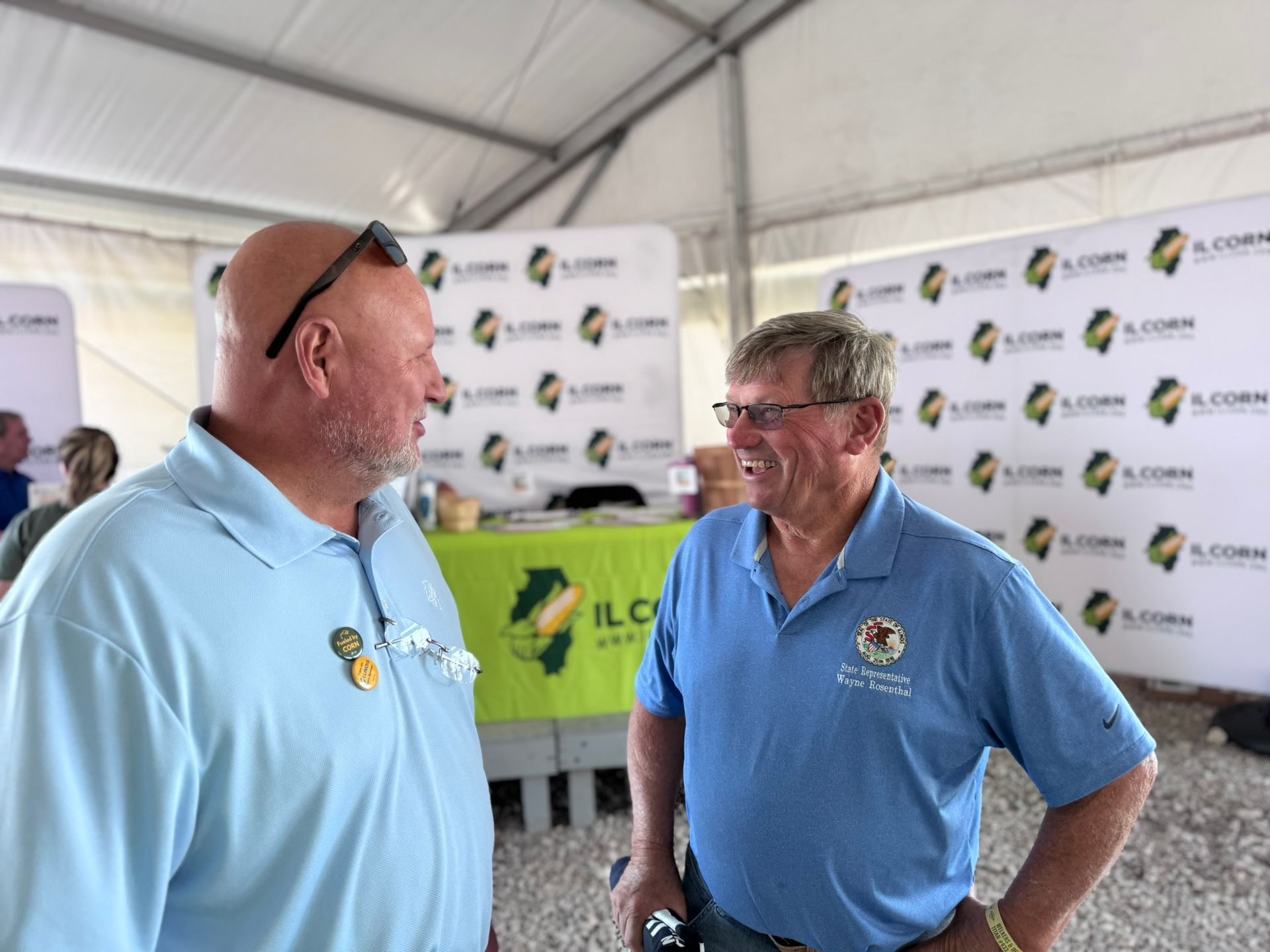 Two men in blue shirts converse under a tent with 