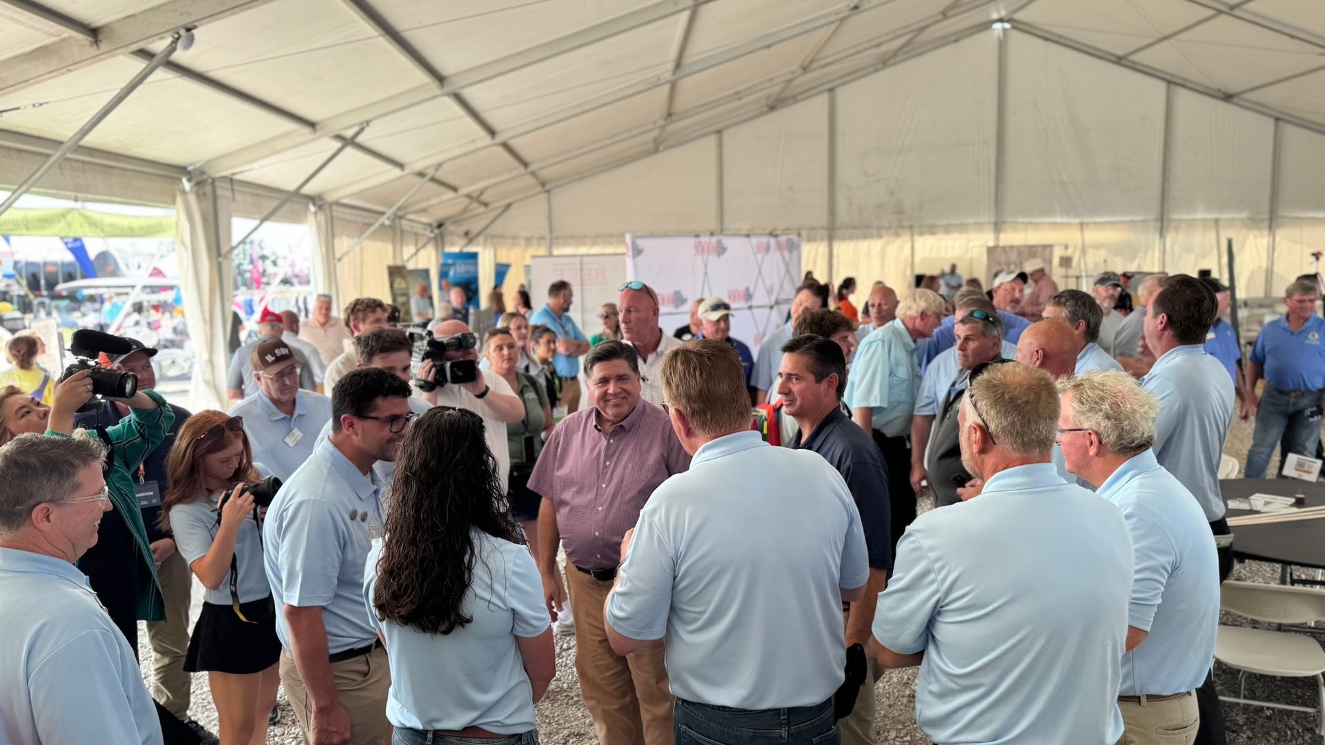 Group of people gathered inside a tent, talking. Men mostly, wearing light blue shirts, talking and smiling.