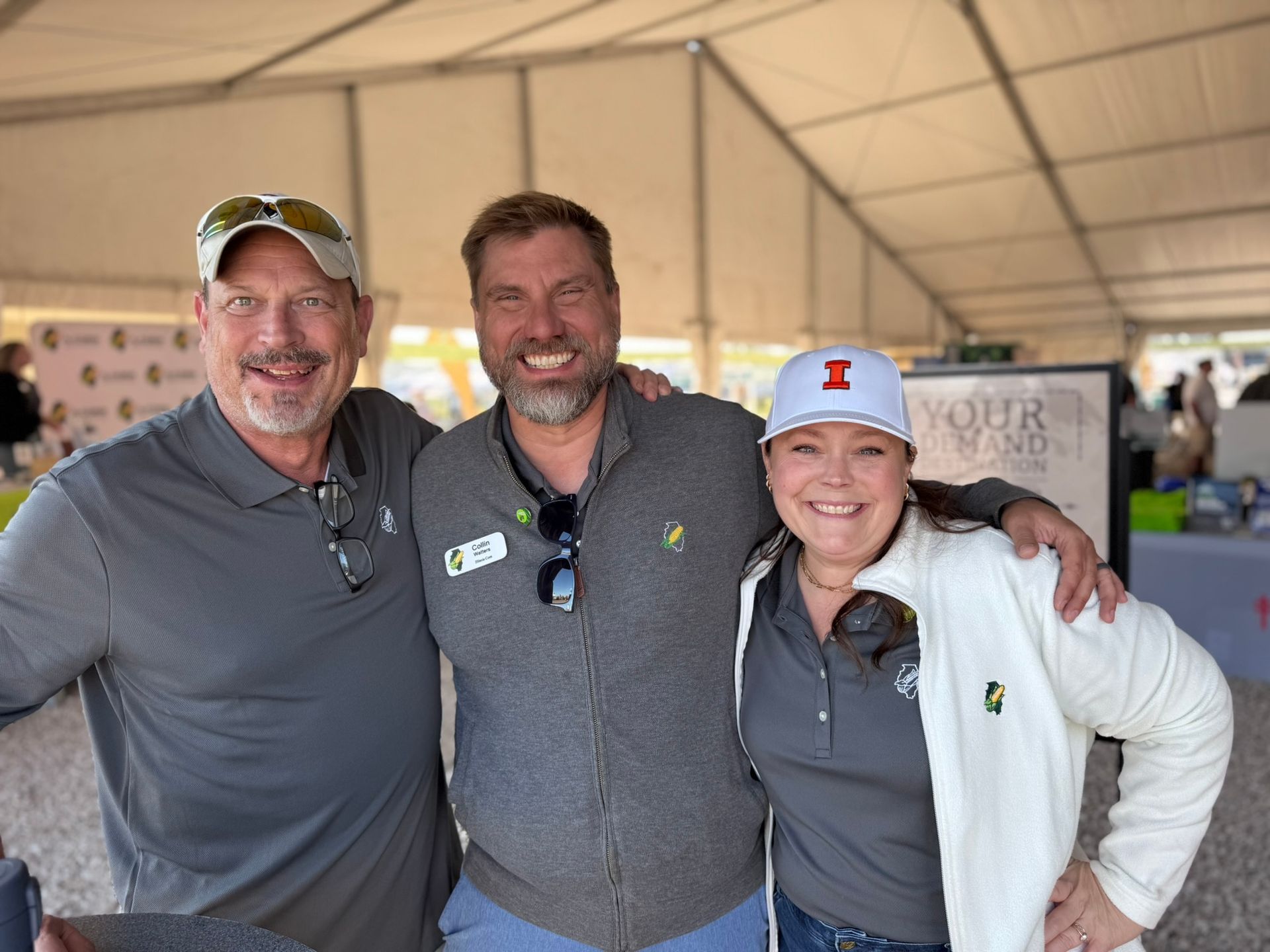 Three smiling people pose outdoors under a tent; two men and a woman; one in a white cap.