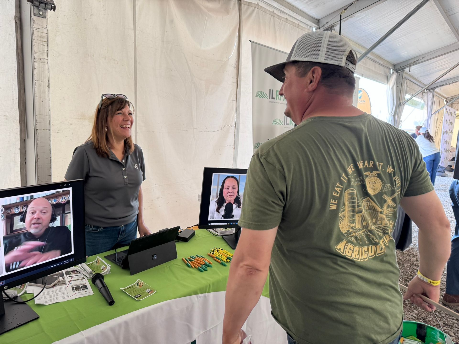 Woman at a booth talks to a man in a green shirt. Video calls appear on monitors. Outdoor setting.