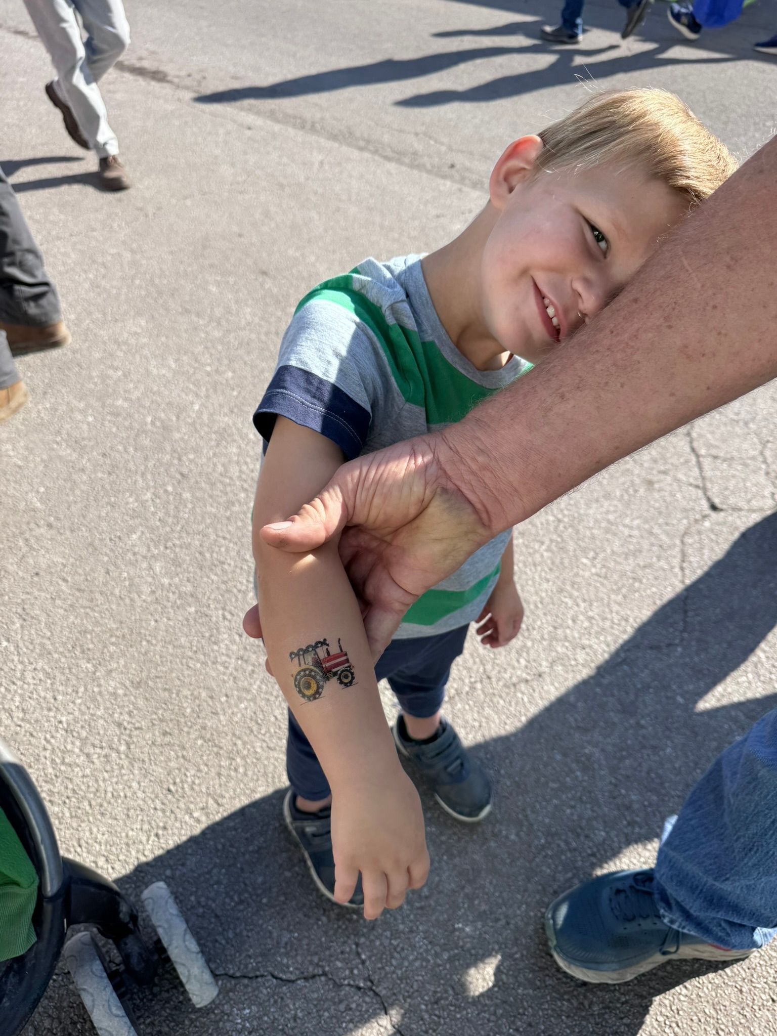 Young boy smiles showing temporary tattoo of a combine on his arm. Outdoors, gray pavement.