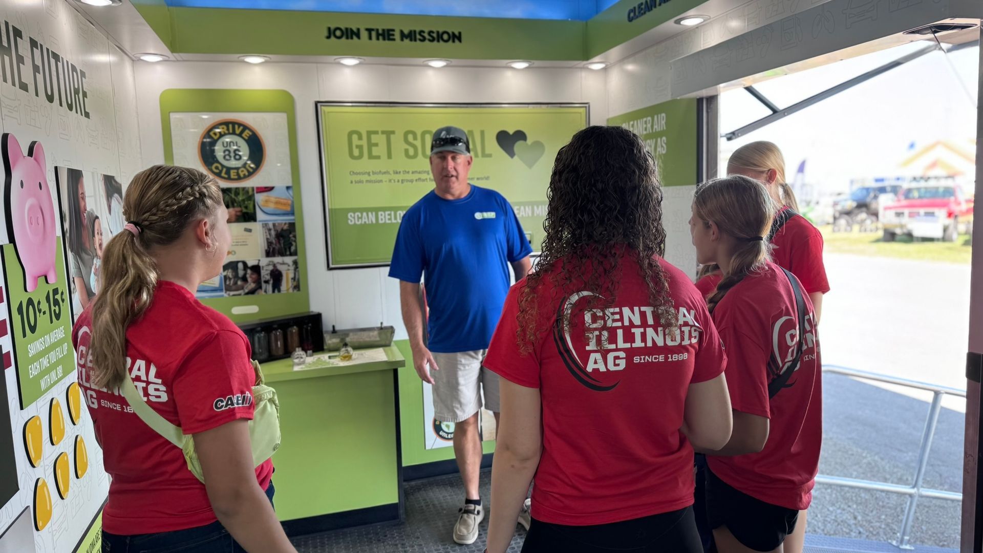 People at an agriculture event, with a man in a booth surrounded by young people in red shirts.
