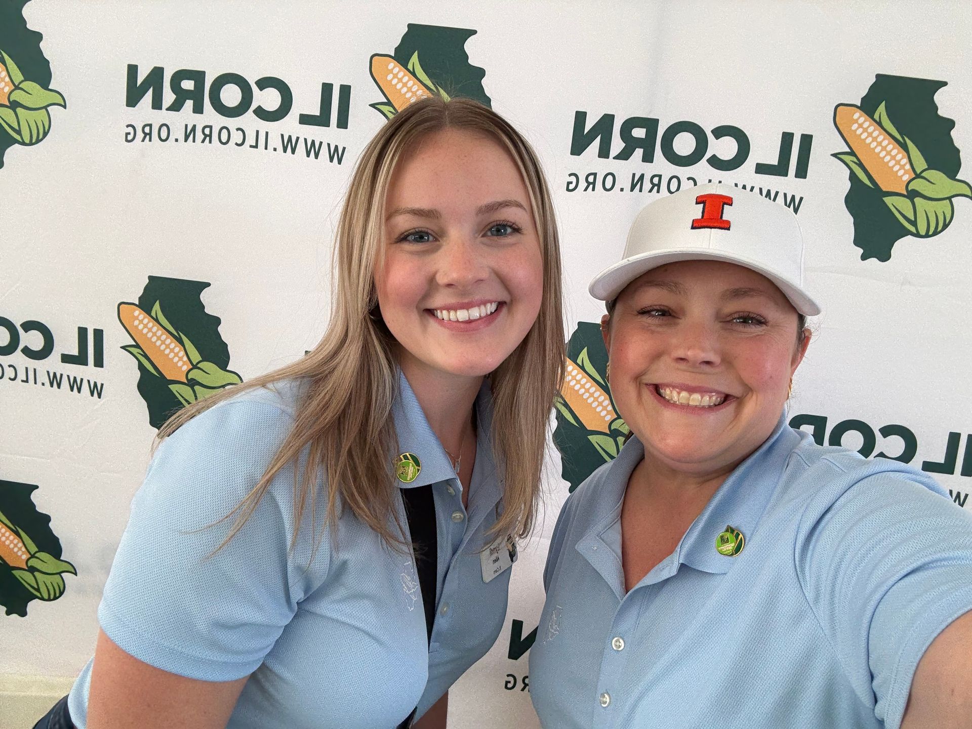 Two women smiling, wearing light blue shirts with a corn logo, posing in front of a backdrop with corn graphics.