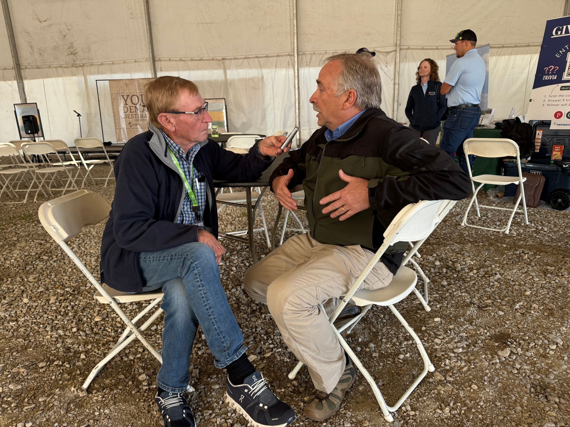 Two men in a tent, one interviewing the other. They sit in chairs, gesturing. Others stand nearby.