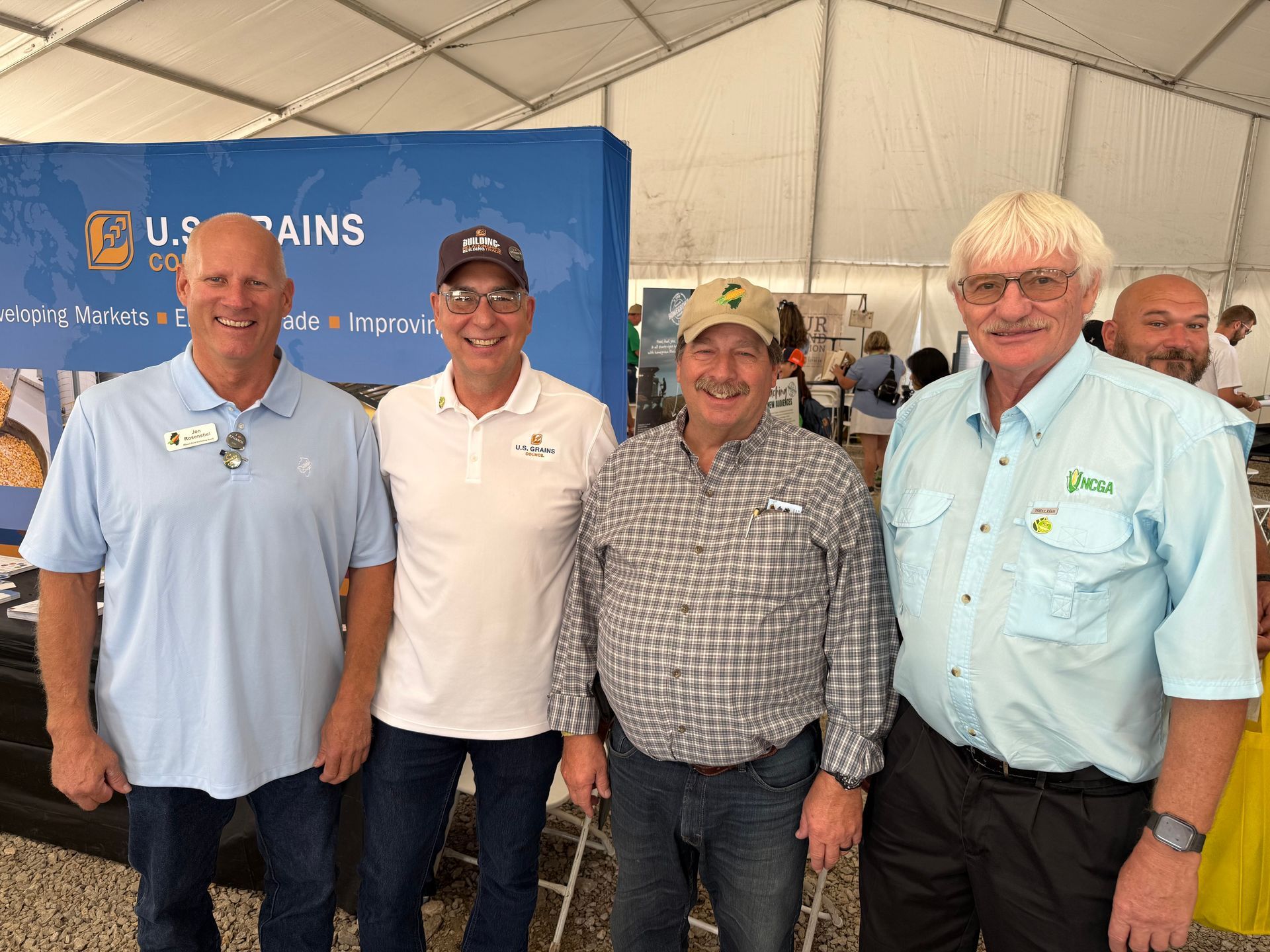 Four men in shirtsleeves pose in front of a banner at an outdoor event.