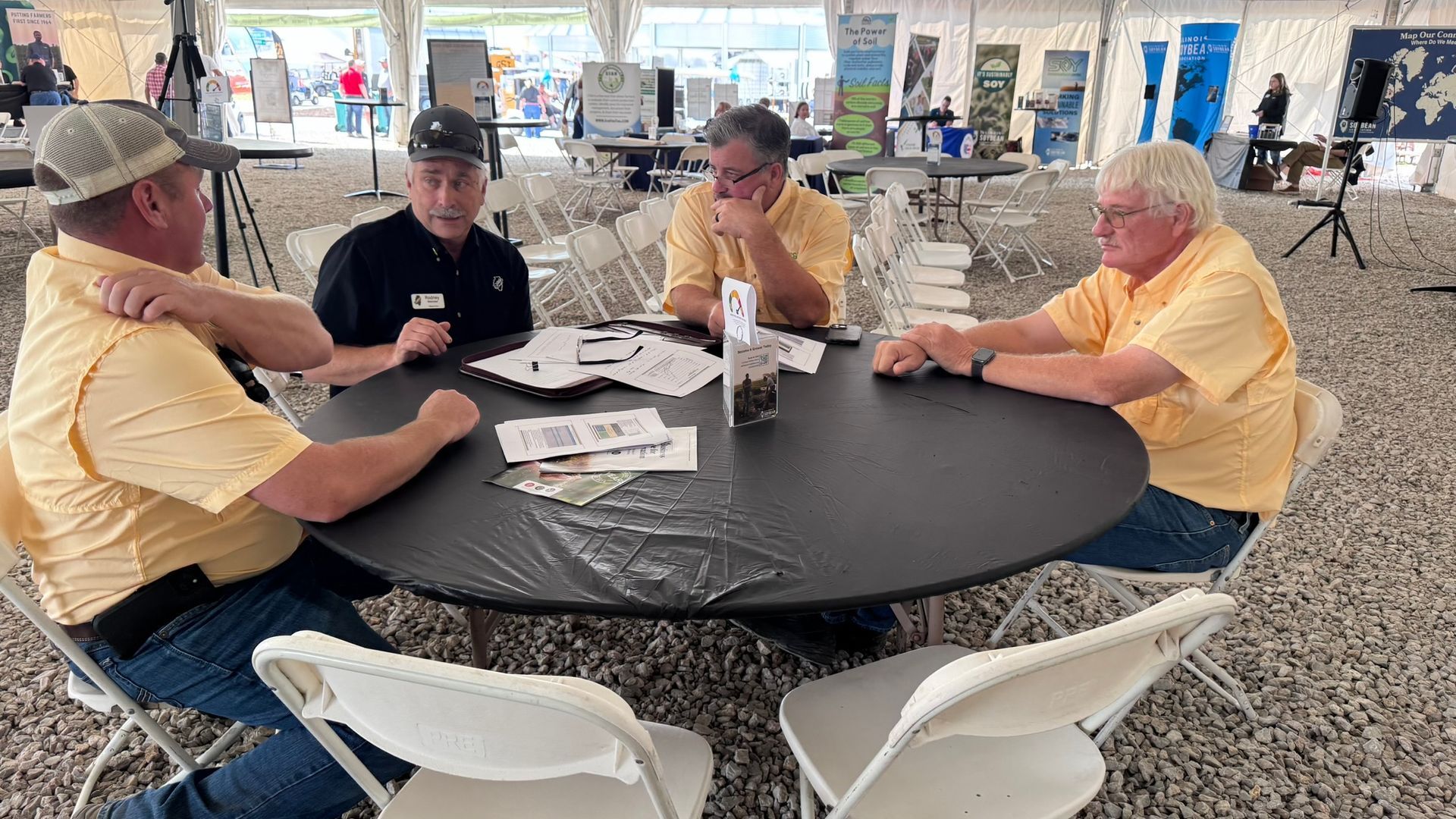 Four men in yellow shirts seated around a black table, discussing papers inside a tent.