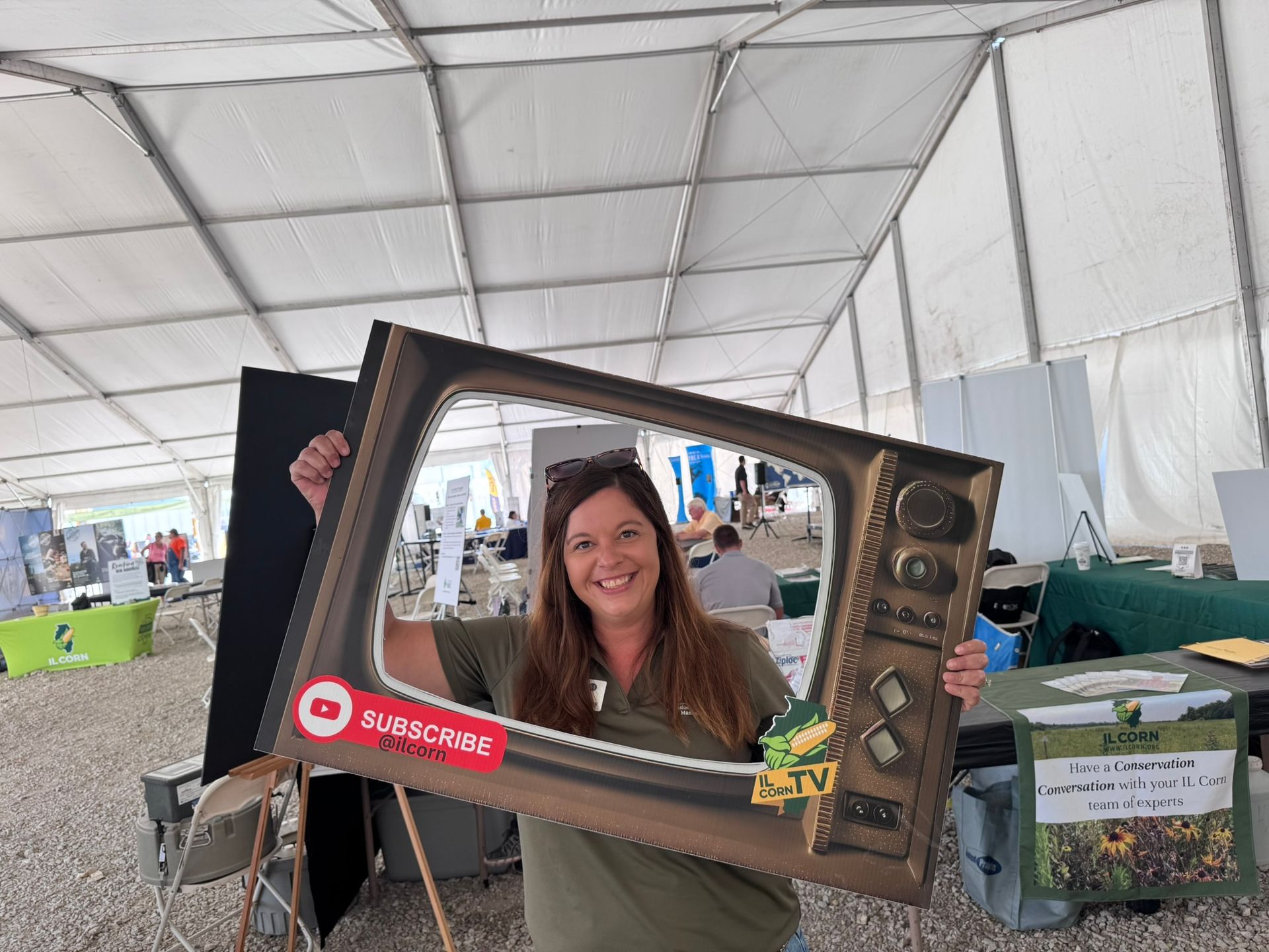Woman holding a cardboard TV frame with YouTube subscribe button, smiling outdoors. Tent setting, various booths visible.