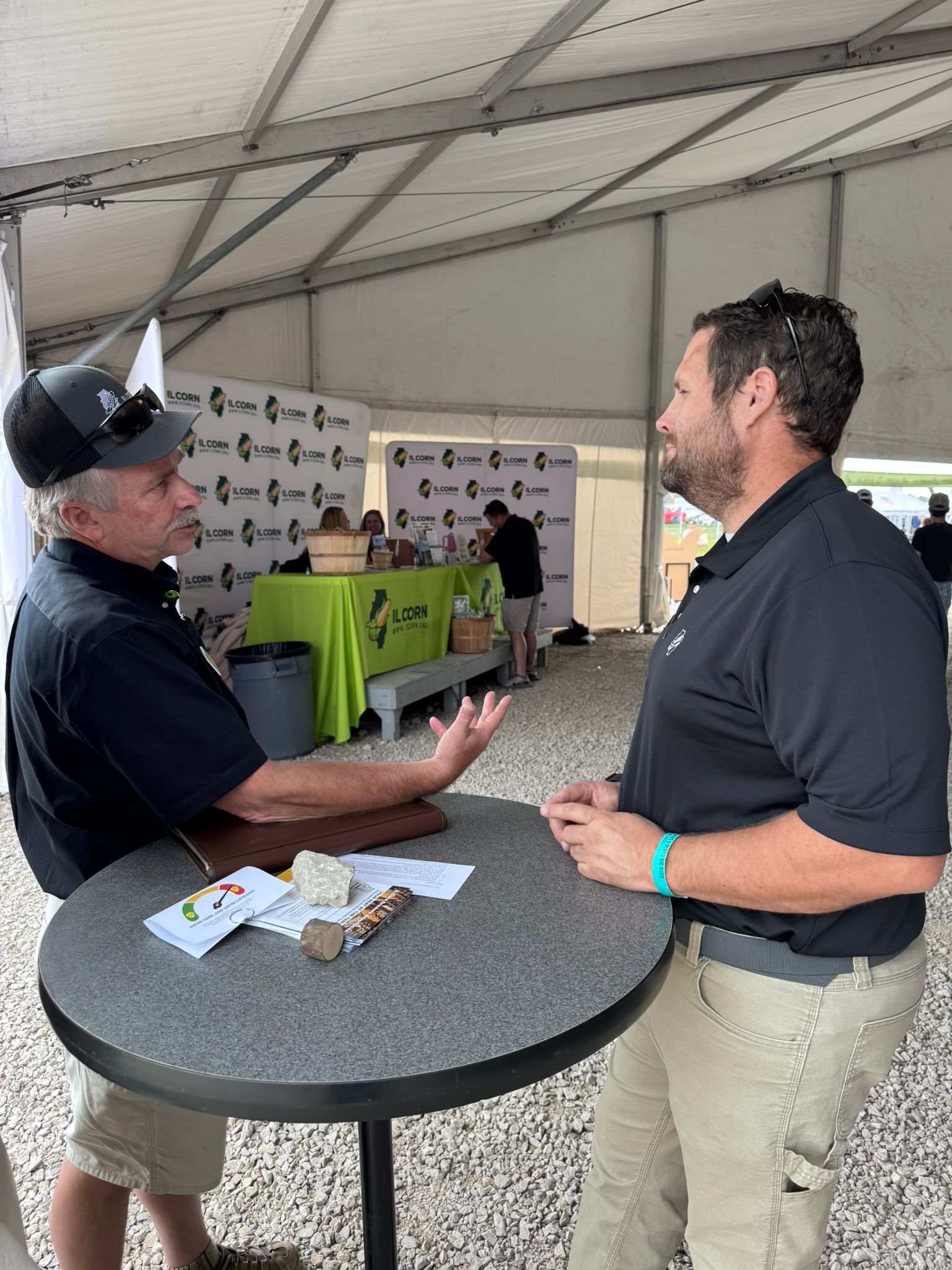Two men converse at a small table under a tent. One gestures with his hand, the other listens.