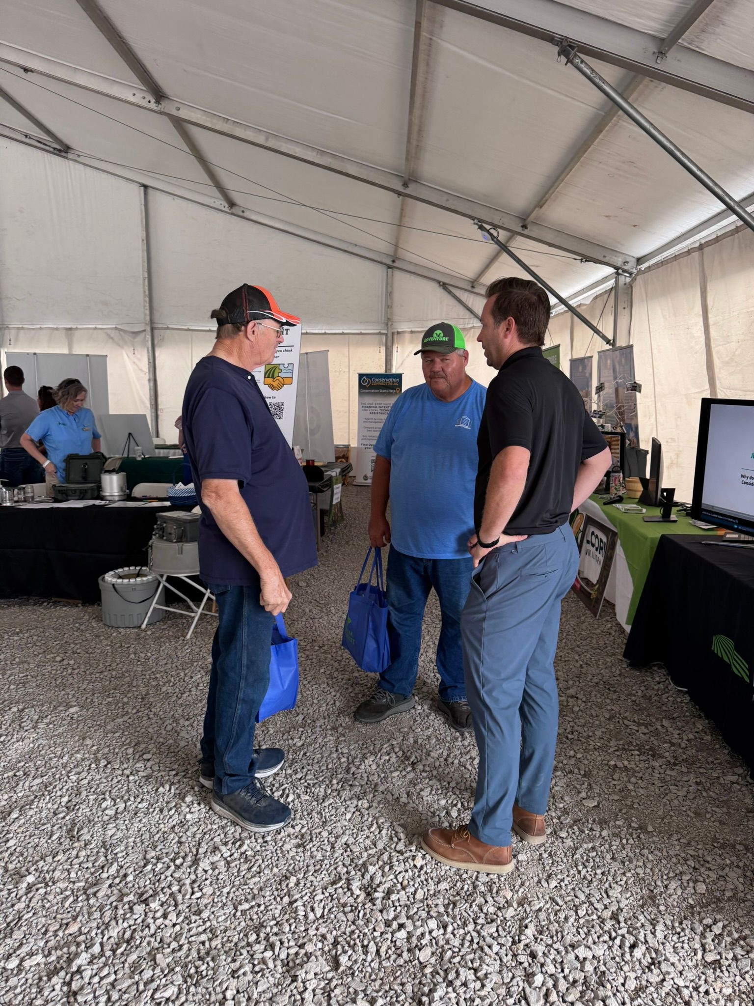 Three men converse inside a tent at an event. They are standing near display tables.