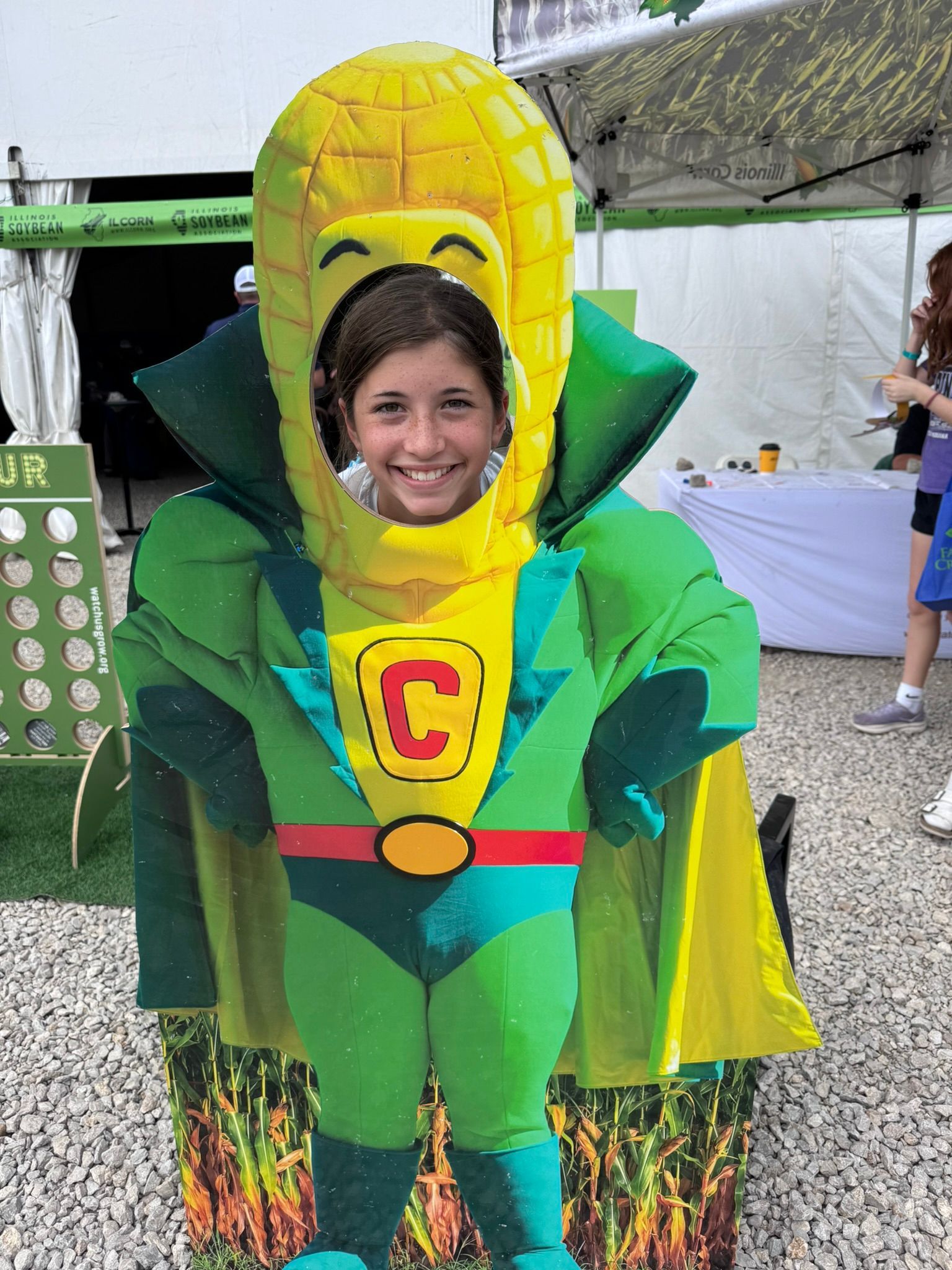Girl poses as Captain Corn cutout at an outdoor event. She smiles, wearing a green suit.