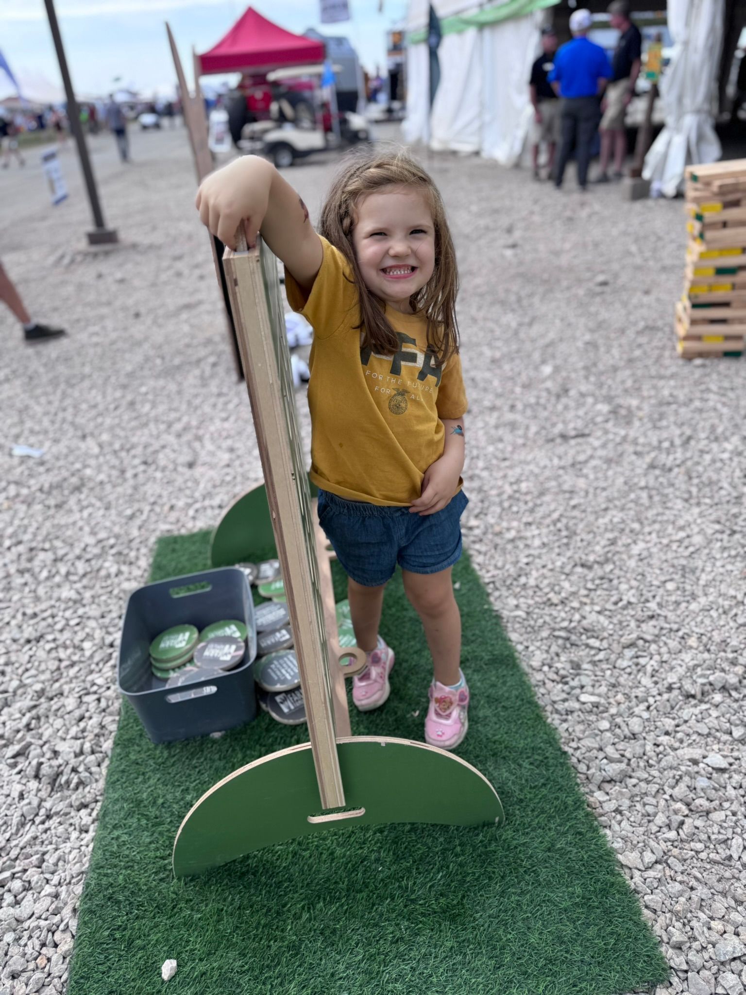 Young girl with curly hair smiles while holding a lawn game in a fair setting.