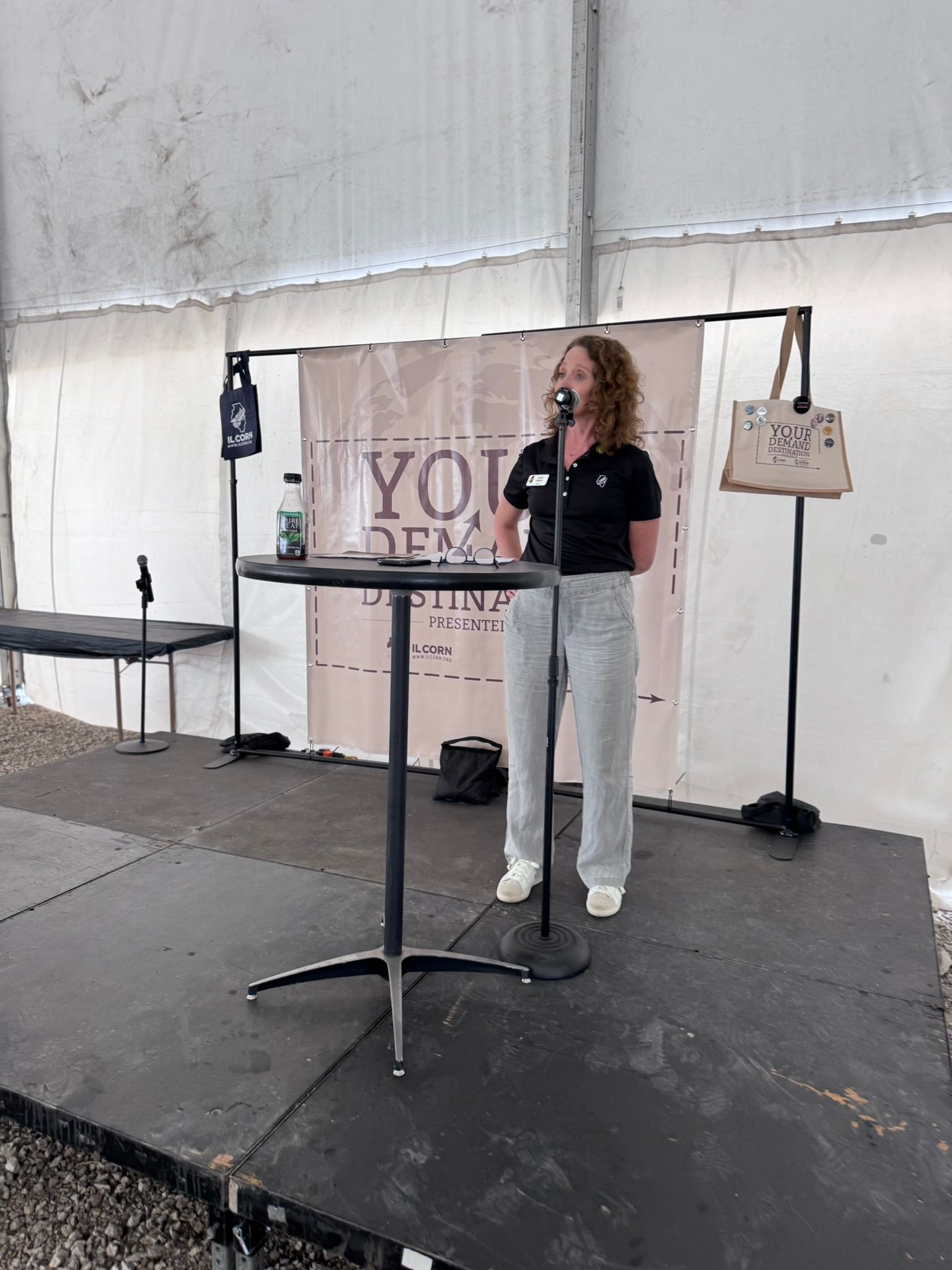 Woman speaking at a podium on a stage, wearing a black shirt and gray pants, inside a tent.