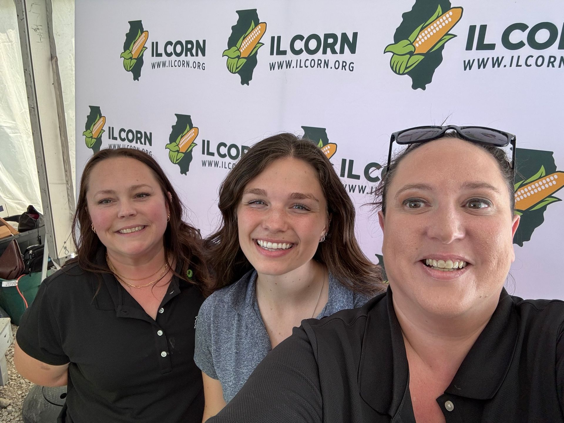 Three women smile in front of an IL Corn banner.