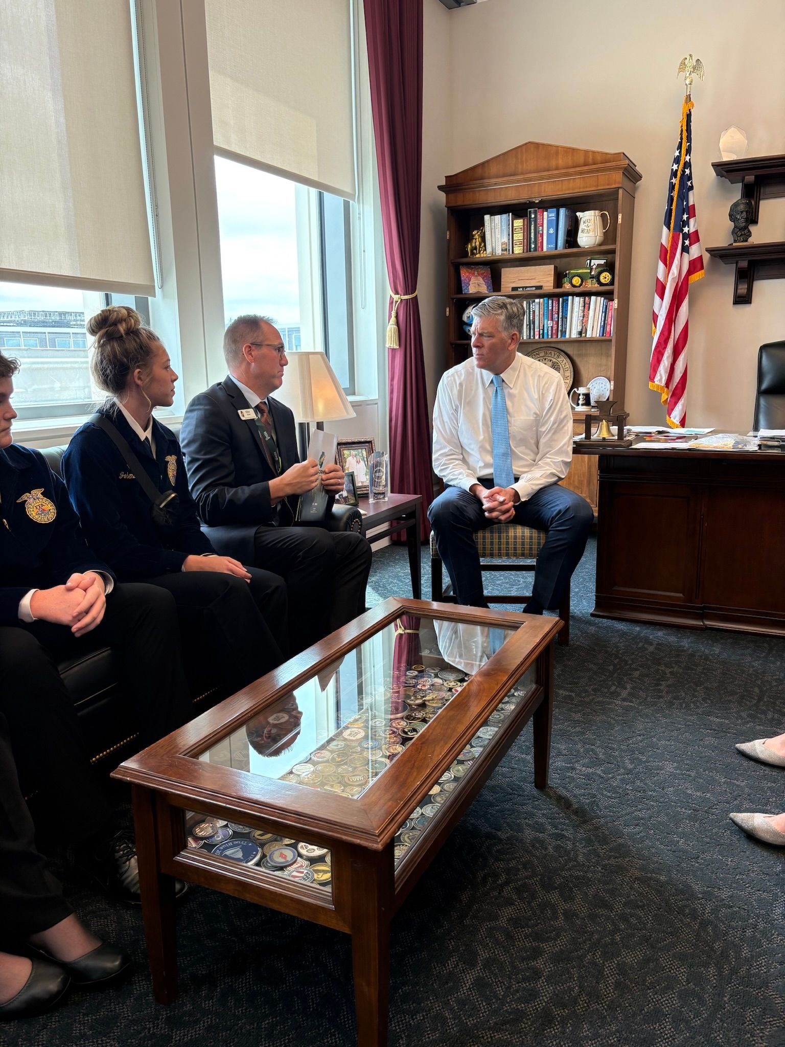 People in a room, one man is speaking, and a group of people are listening. American flag and desk are present.