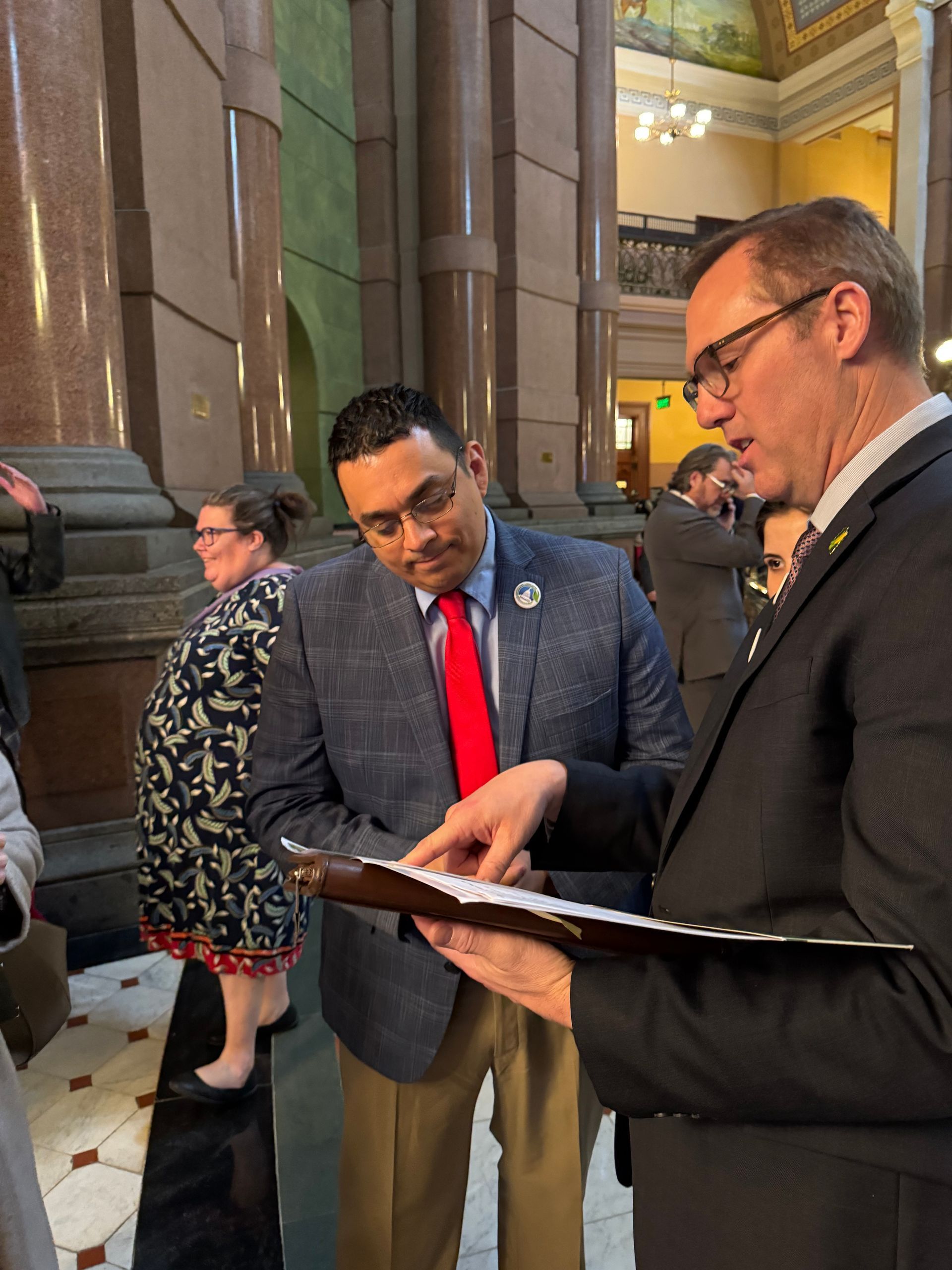Two men reviewing papers in a government building. One points at the document; a third person is visible.