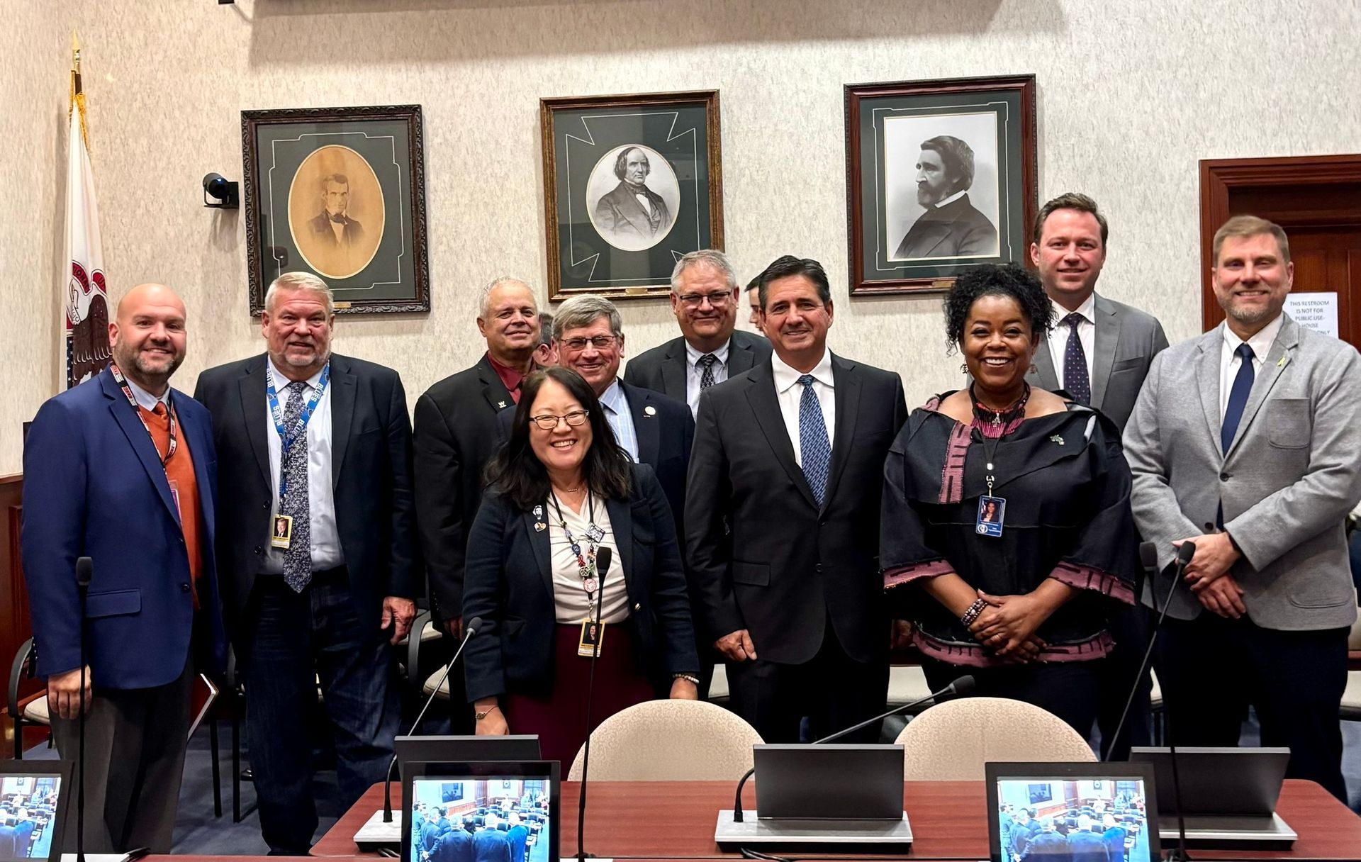 A panel of people in a meeting room, facing each other at a long table. A video camera is set up in front.