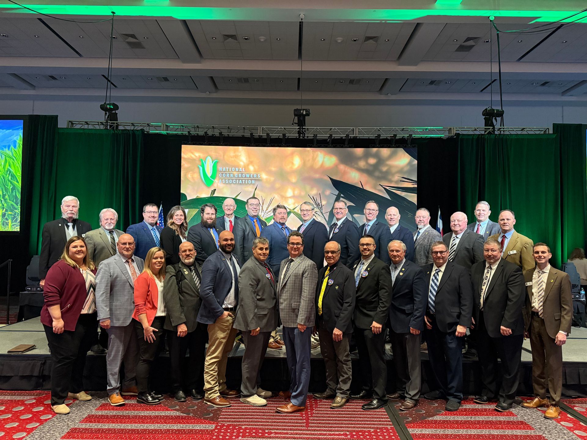 Group of people posing on a stage, with a green backdrop. Some are wearing suits, smiling.