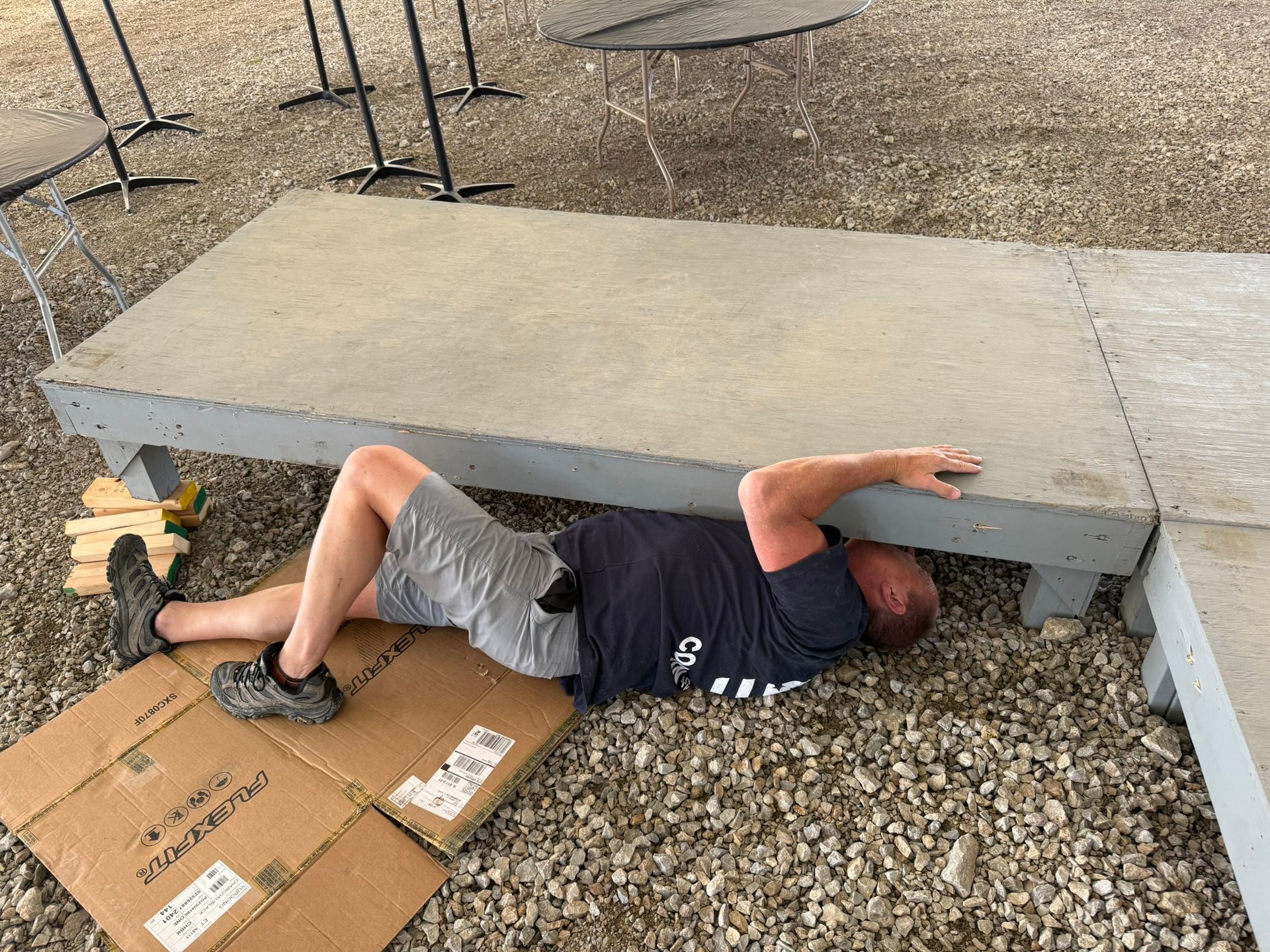 Man lying under a gray platform outdoors, wearing shorts and a t-shirt.