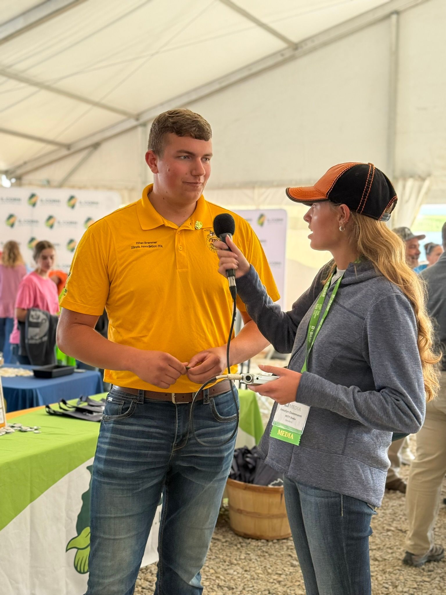 Man in yellow shirt being interviewed by a woman holding a microphone, at an outdoor event.