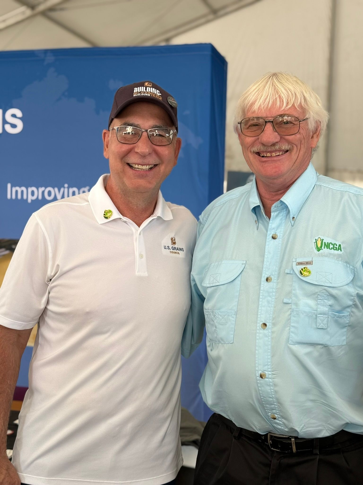 Two men smiling for a photo. One wears a hat and white shirt, the other, a light blue shirt with logo. Behind them, a blue banner.