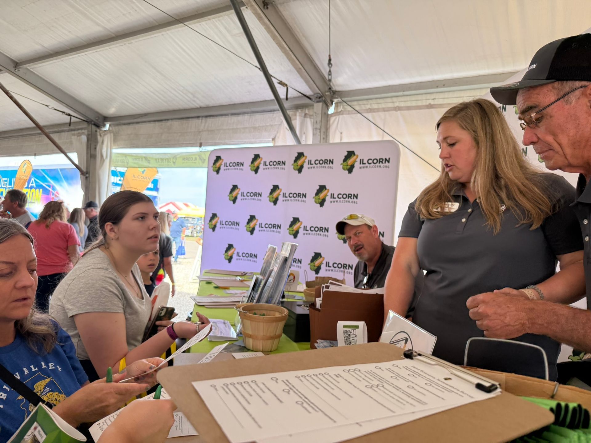 People at an outdoor event booth, interacting with staff. Signage and promotional materials on display.