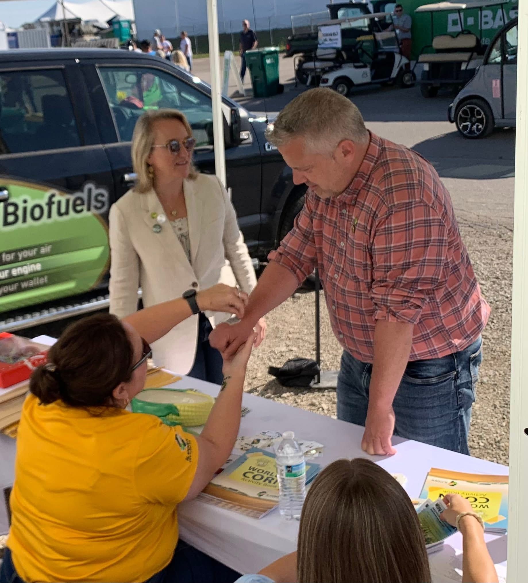 Man and woman at booth, man's arm examined. Others watch. Outdoor setting.