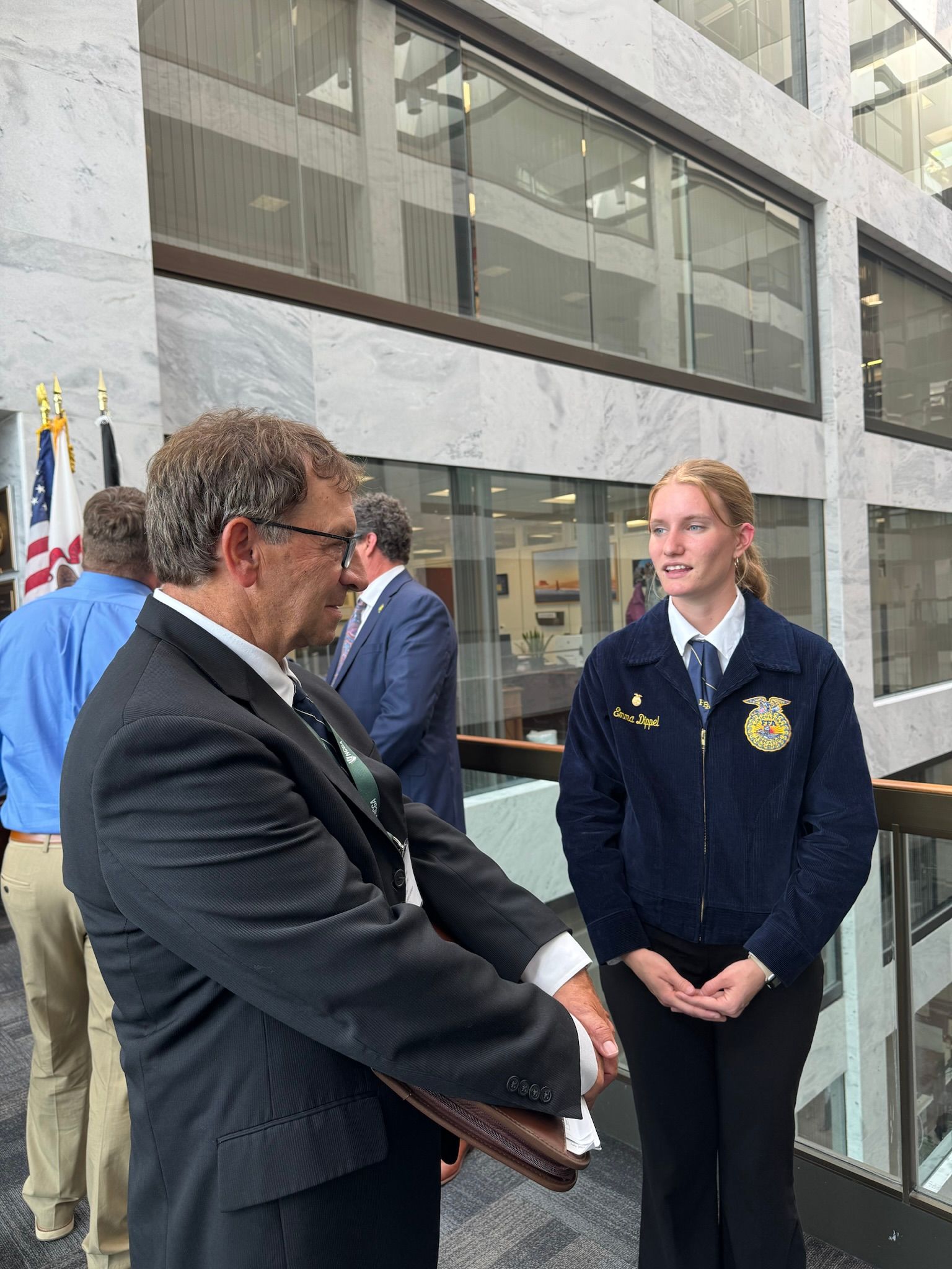 Man in suit presents a document to a young woman in FFA jacket, inside a building.