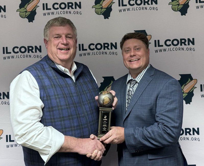 Two men shaking hands in front of a wall that says il corn