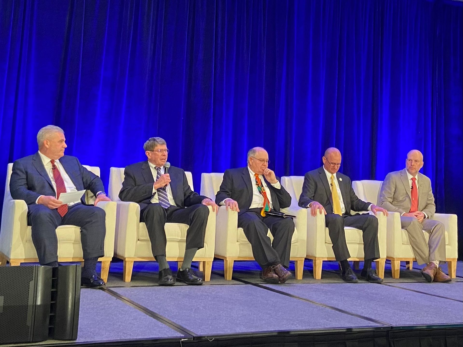 A group of men in suits and ties are sitting in chairs on a stage.