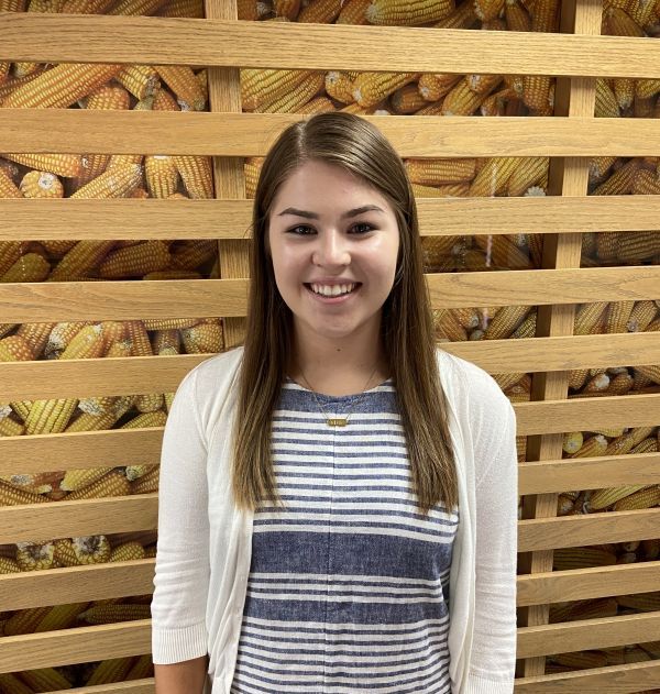 A woman is standing in front of a wooden wall filled with corn.
