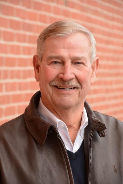 A man wearing a brown leather jacket and a white shirt is smiling in front of a brick wall.