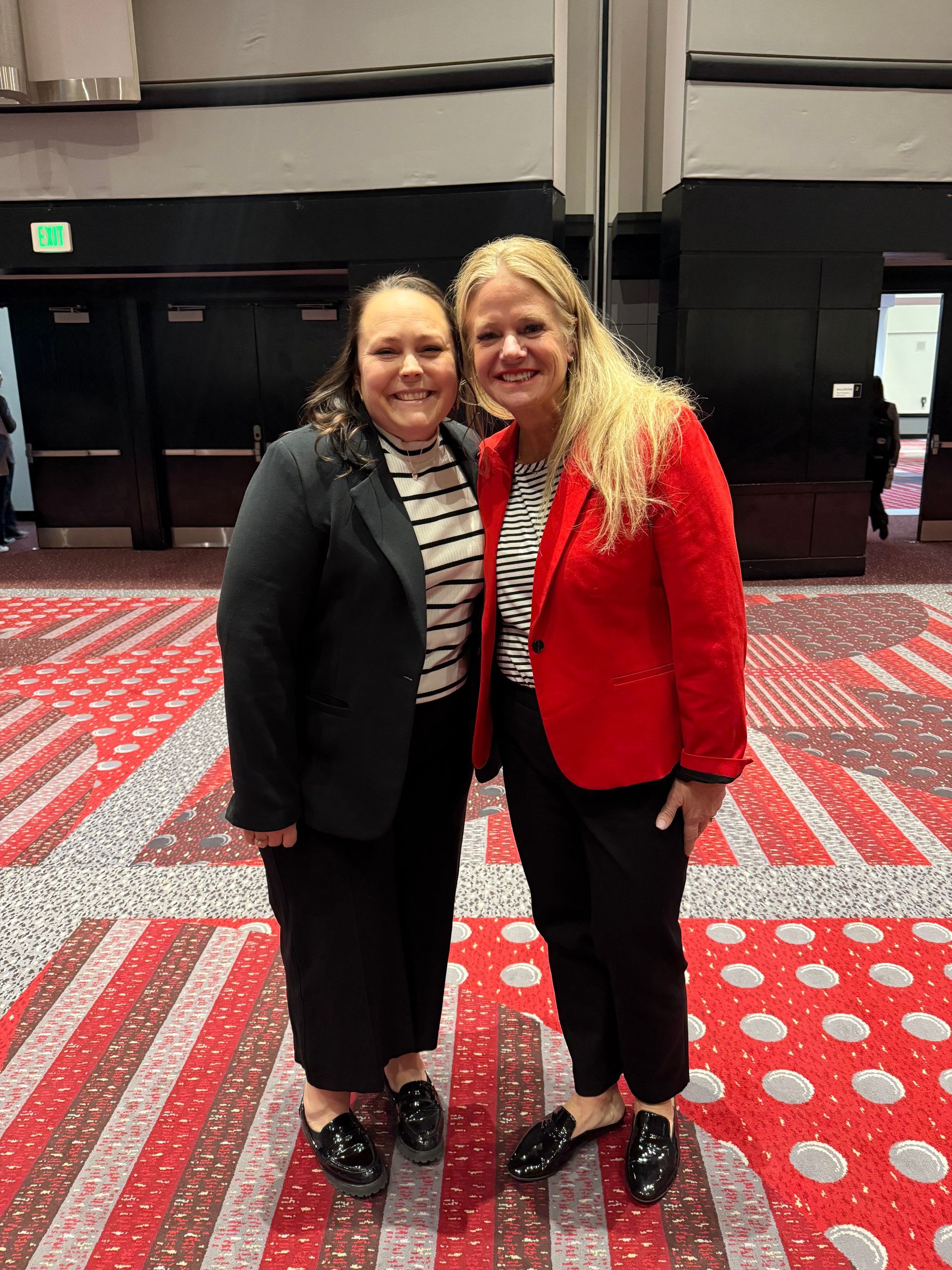 Two women smiling, posing for a photo. One in a red blazer, the other in a black blazer. Striped shirts, black pants, patterned floor.