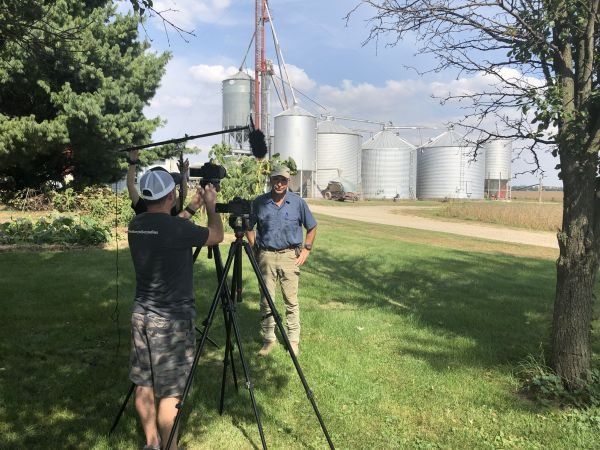 A man is standing in the grass talking to a cameraman.