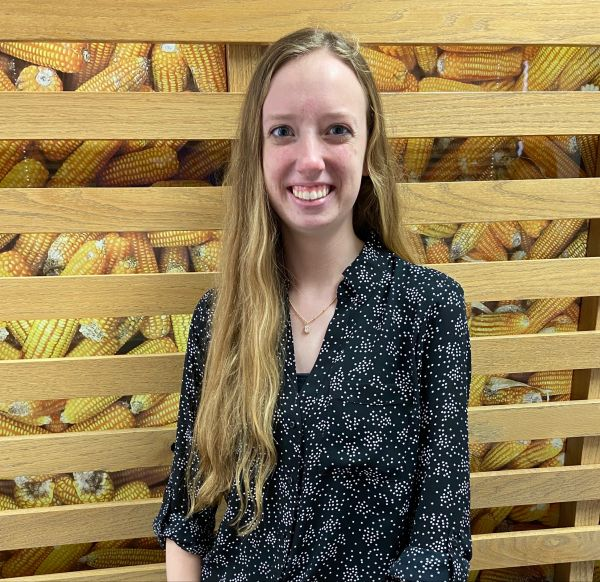 A woman is smiling in front of a wall of corn on the cob